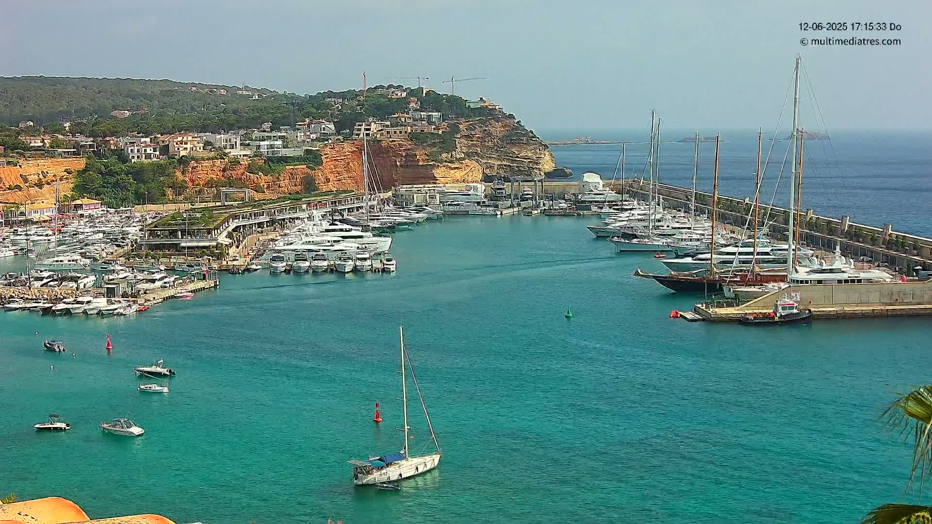 A marina filled with numerous yachts and boats is situated in a calm, turquoise body of water, with a hilly, residential area visible in the background under a clear sky.