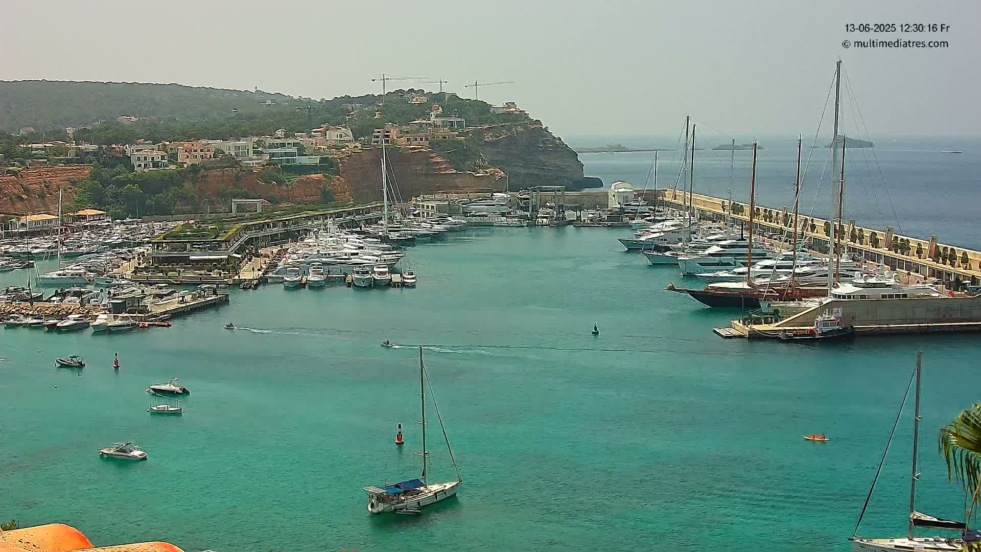 A marina filled with numerous boats and yachts is situated in a calm, teal-colored harbor, against a backdrop of a sunny, hilly coastline.