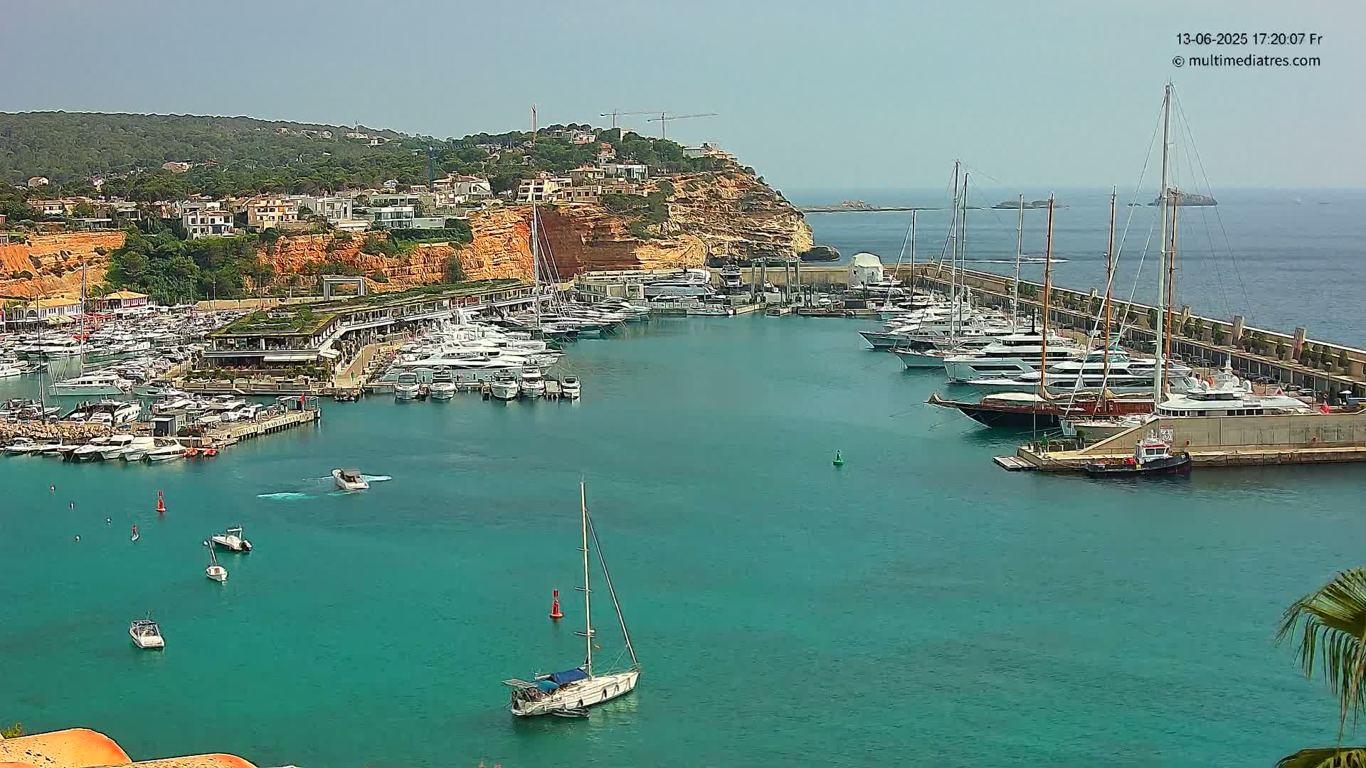 A marina filled with numerous yachts and sailboats is situated along a rocky coastline under a clear, sunny sky.