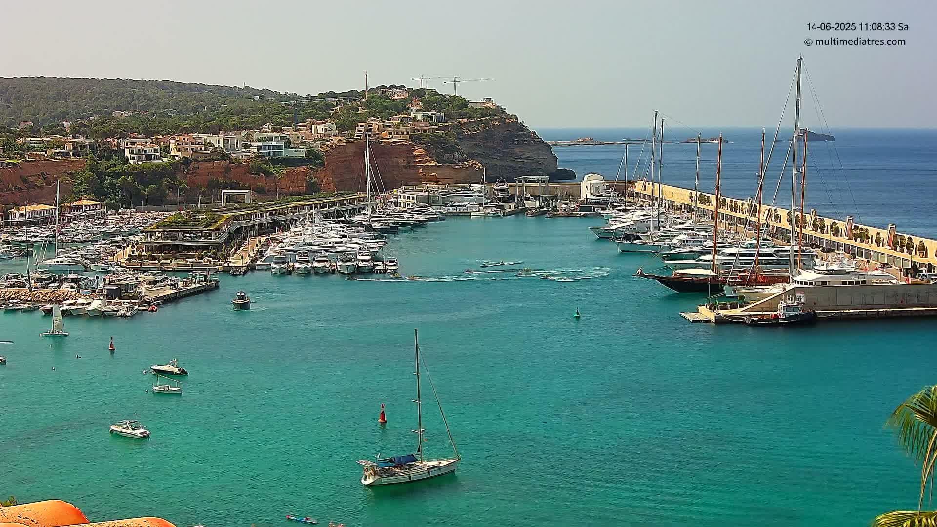 A marina filled with numerous boats and yachts is situated next to a cliffside town on a sunny day.