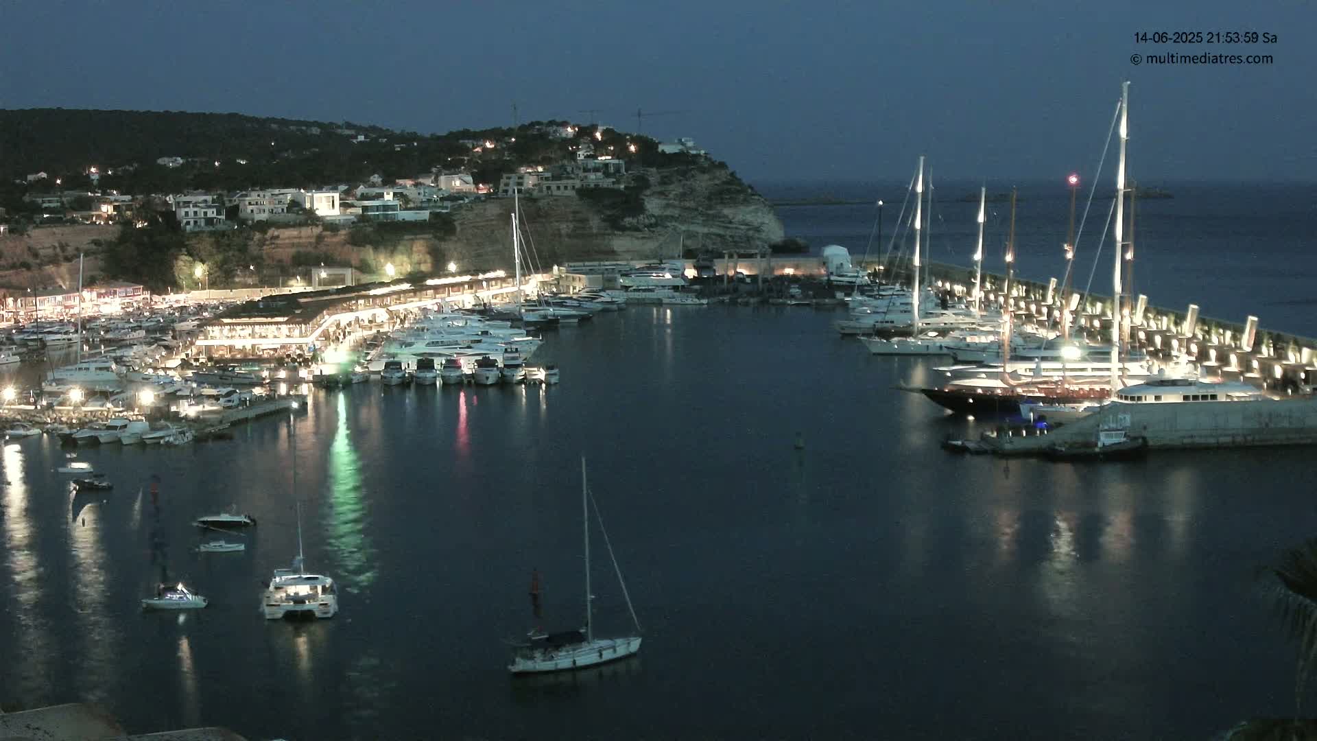 A marina filled with numerous yachts and sailboats is nestled into a coastline of red cliffs and lush greenery under a clear, sunny sky.