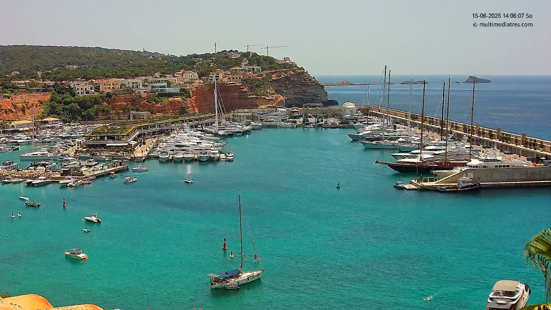 A marina filled with numerous yachts and sailboats is situated in a calm, turquoise bay on a sunny day, with a hilly coastline in the background.