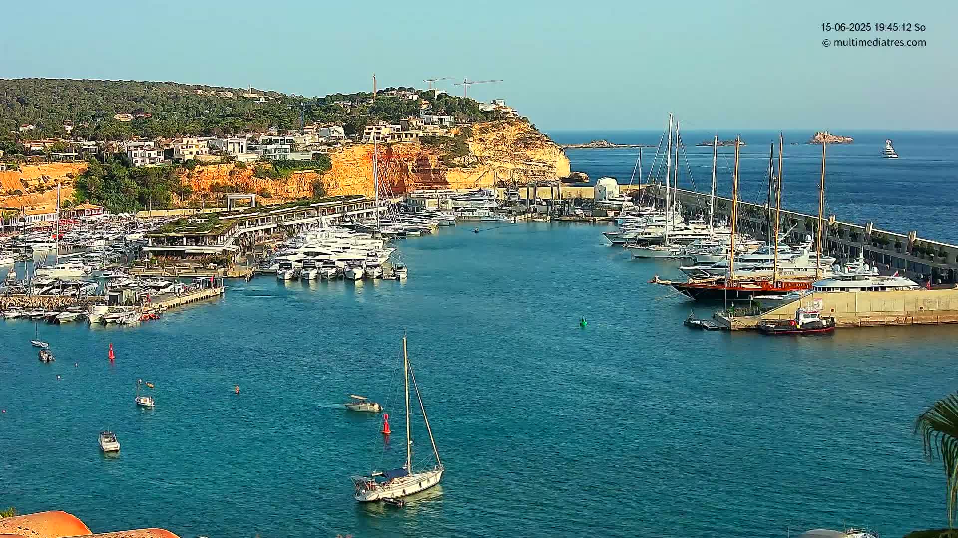 A marina filled with numerous yachts and sailboats is situated on a calm, clear body of water next to a hillside of buildings and vegetation under a sunny sky.