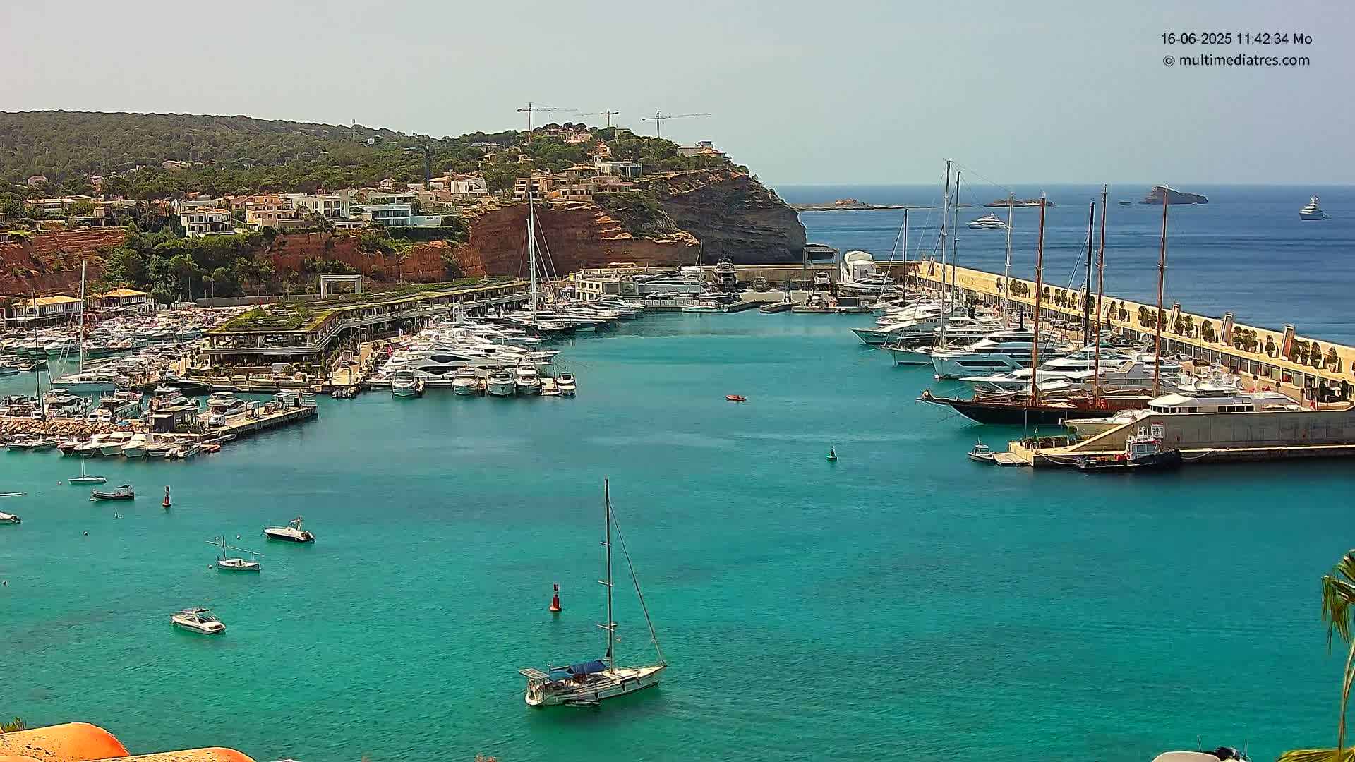 A marina filled with numerous yachts and boats is situated along a rocky coastline on a sunny day, with clear, turquoise water.