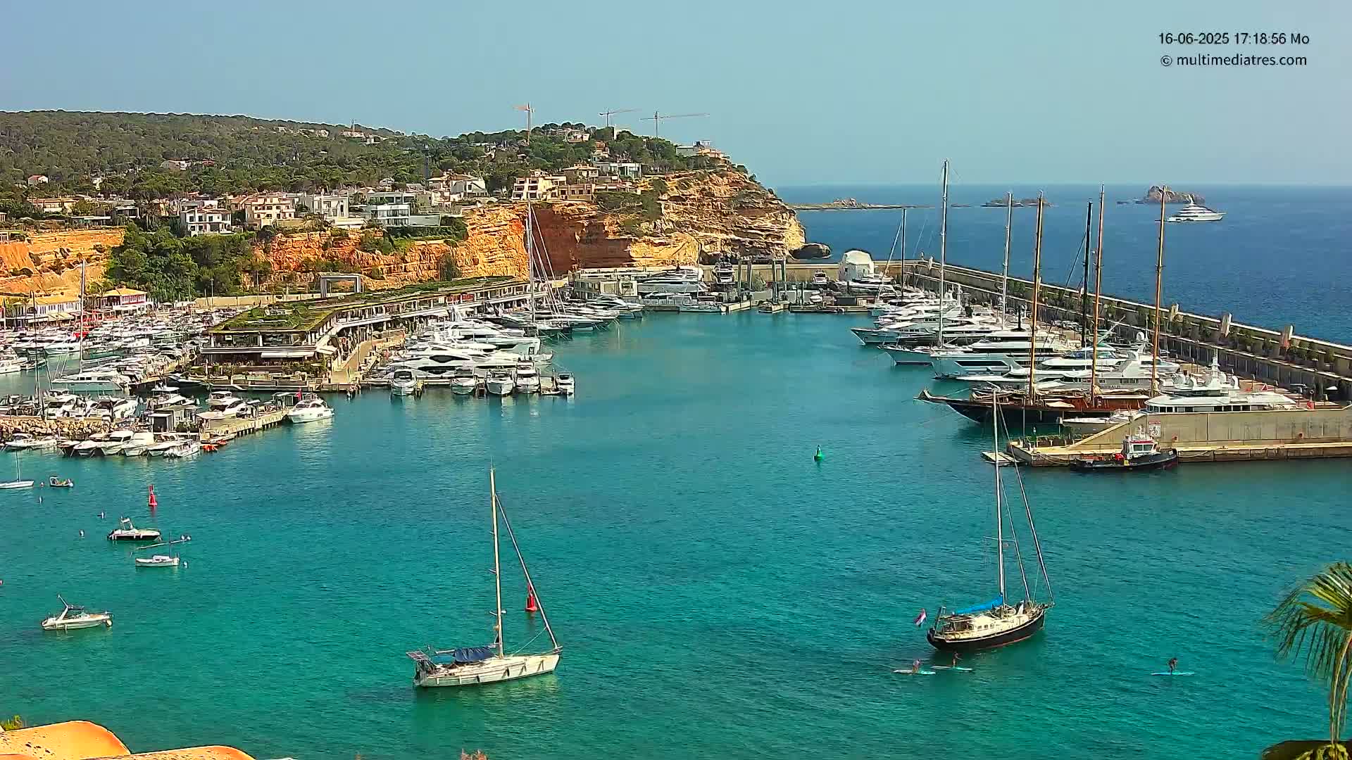 A harbor filled with numerous yachts and sailboats is nestled against a cliffside town under a clear blue sky.