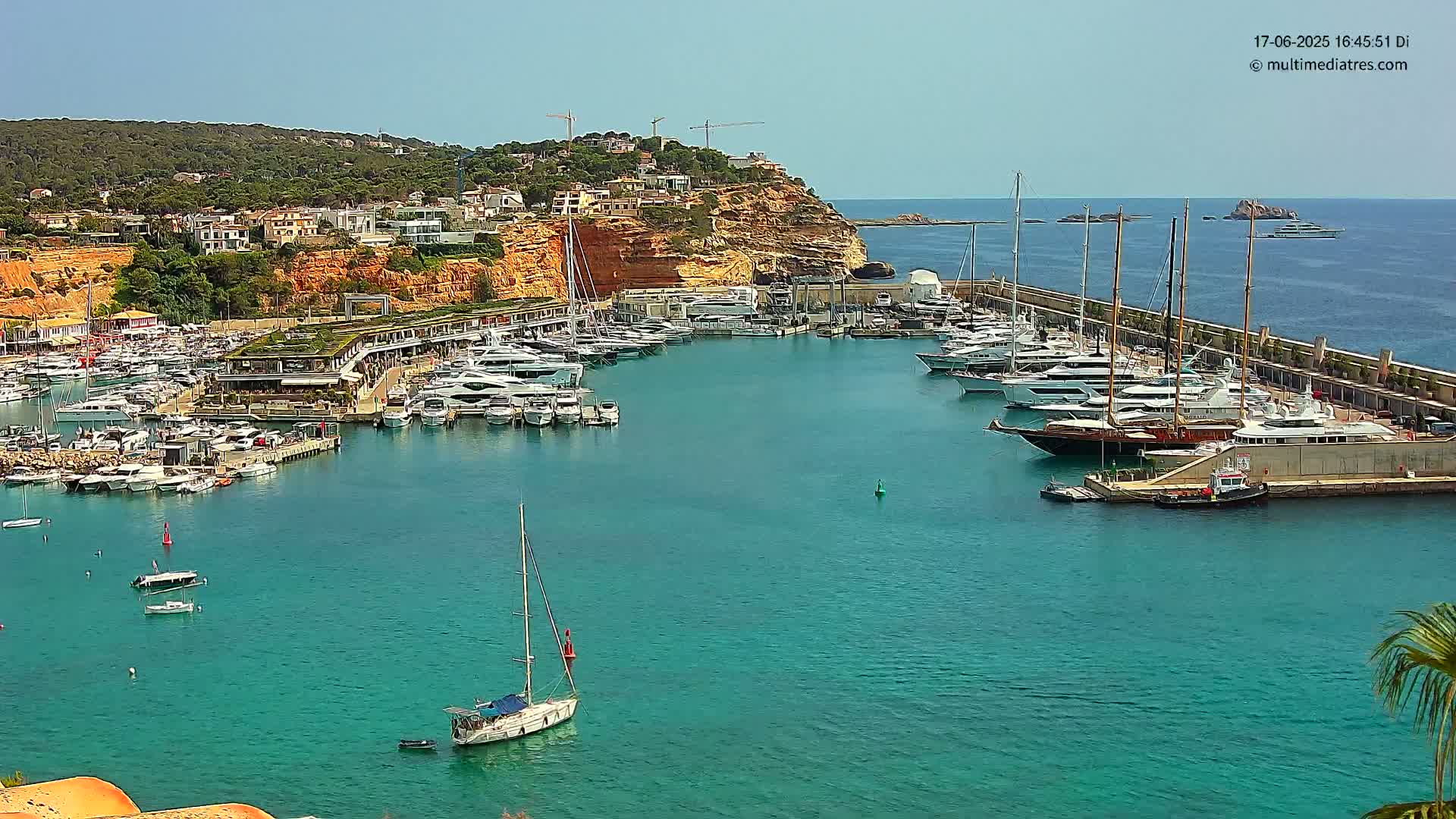 A marina filled with yachts and boats is nestled into a rocky coast under a clear blue sky.