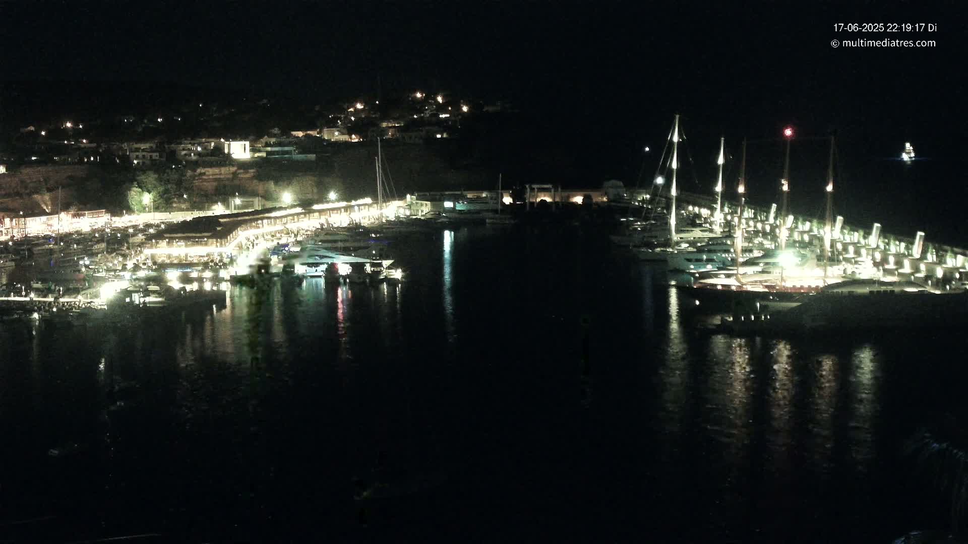 A nighttime view of a harbor filled with boats and yachts, illuminated by lights along the waterfront and on the vessels themselves.