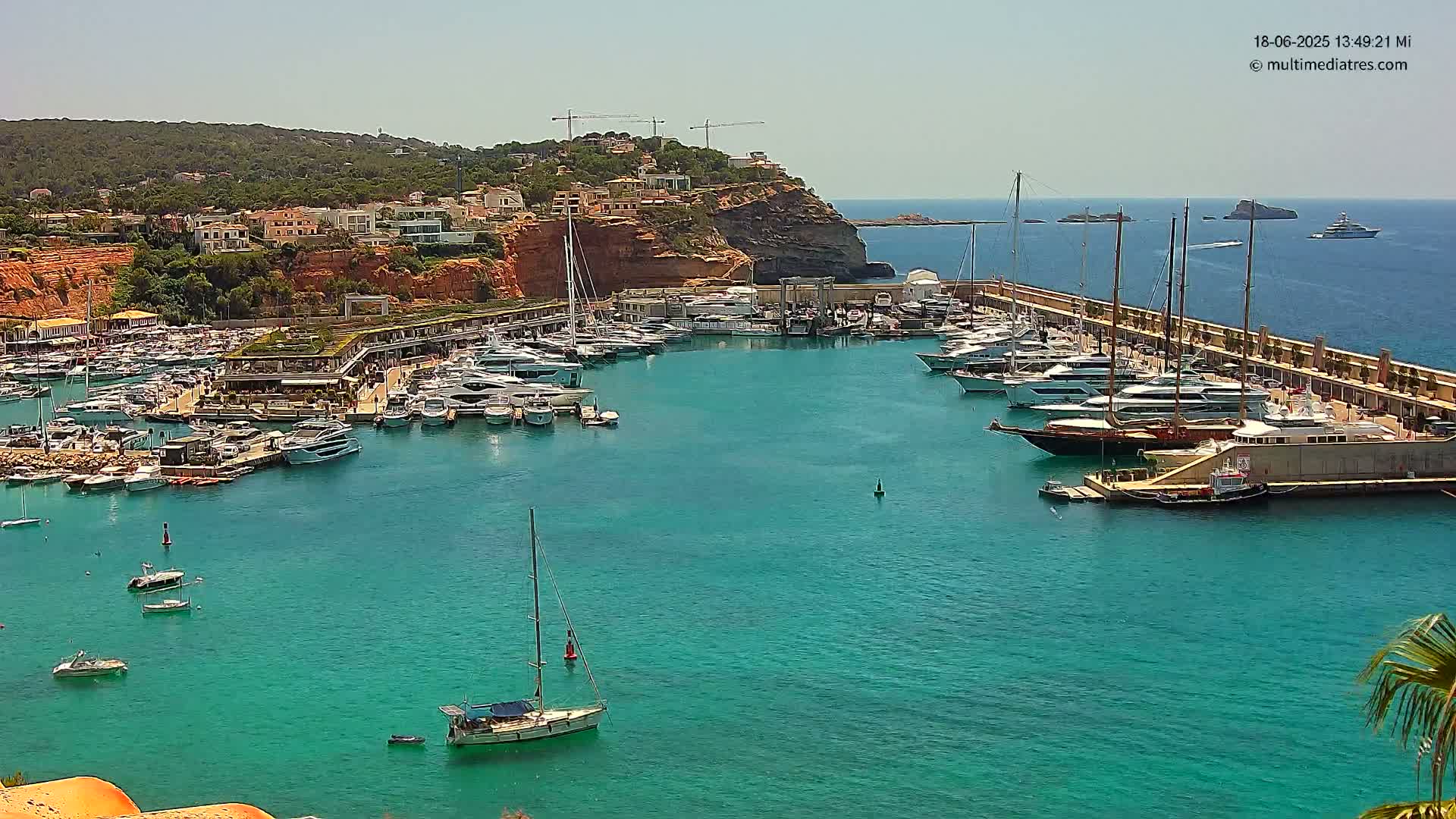 A marina filled with yachts and sailboats is nestled into a turquoise bay on a sunny day, with a hillside of buildings and red cliffs in the background.