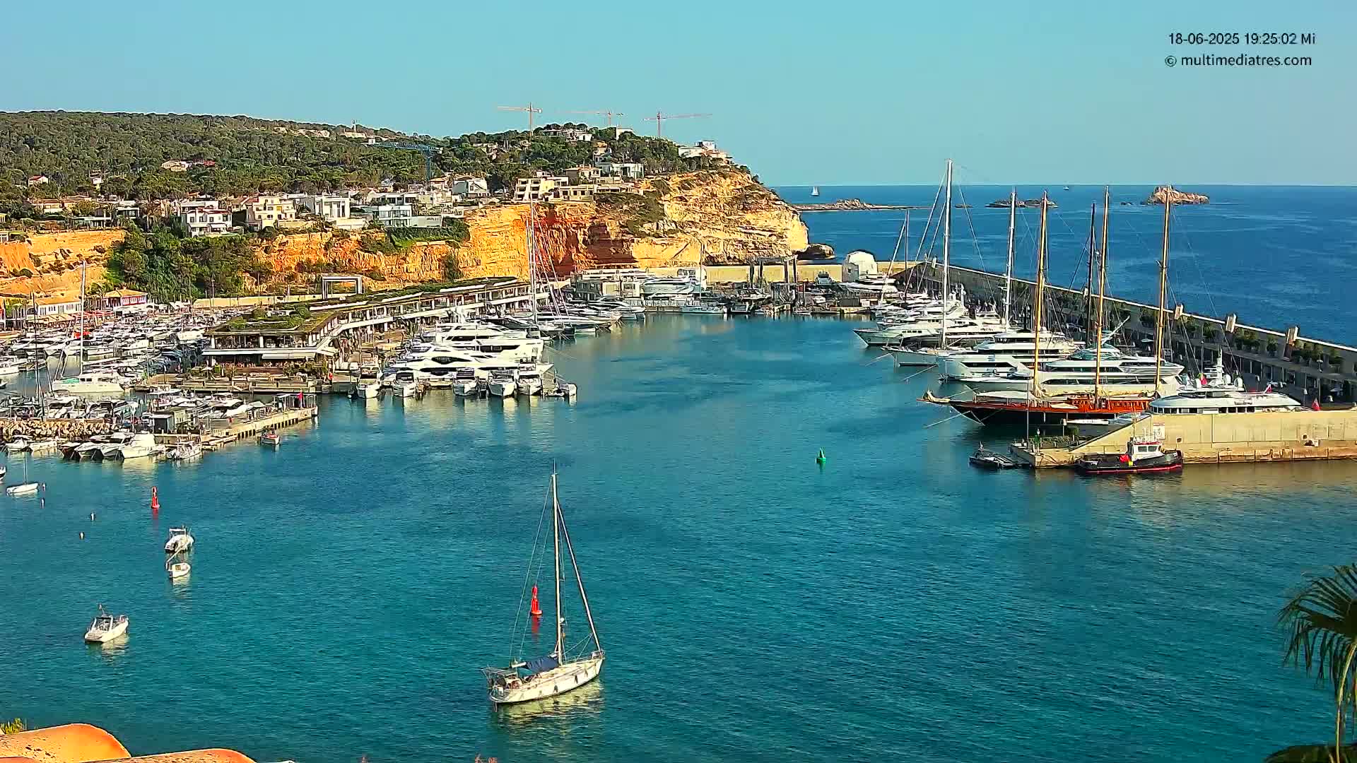 A marina filled with numerous yachts and sailboats is nestled into a coastal area with a clear blue sky and calm water.