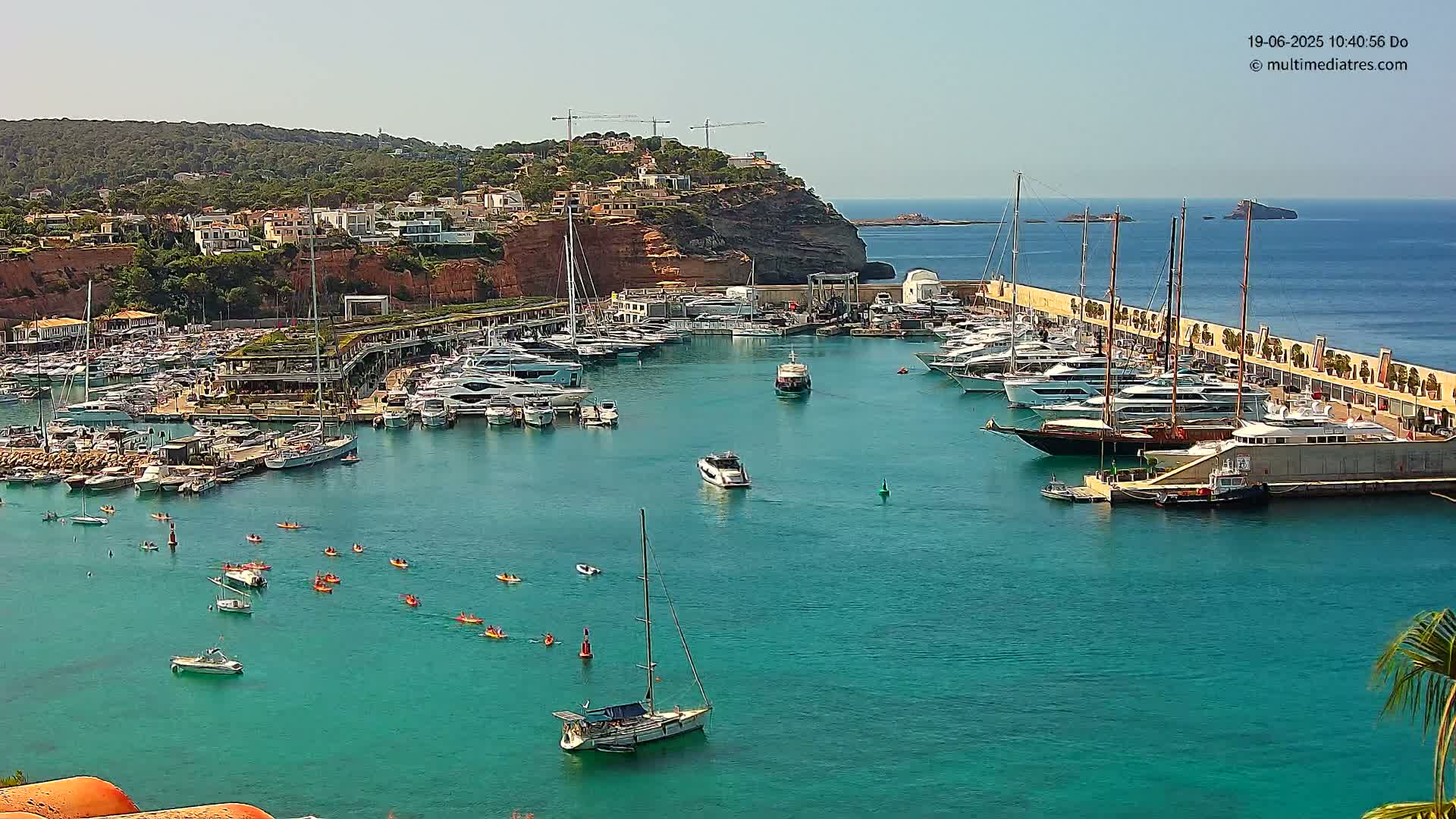 A marina filled with numerous yachts and sailboats, kayakers paddling in the turquoise water, is situated below a hillside of buildings under a bright, sunny sky.