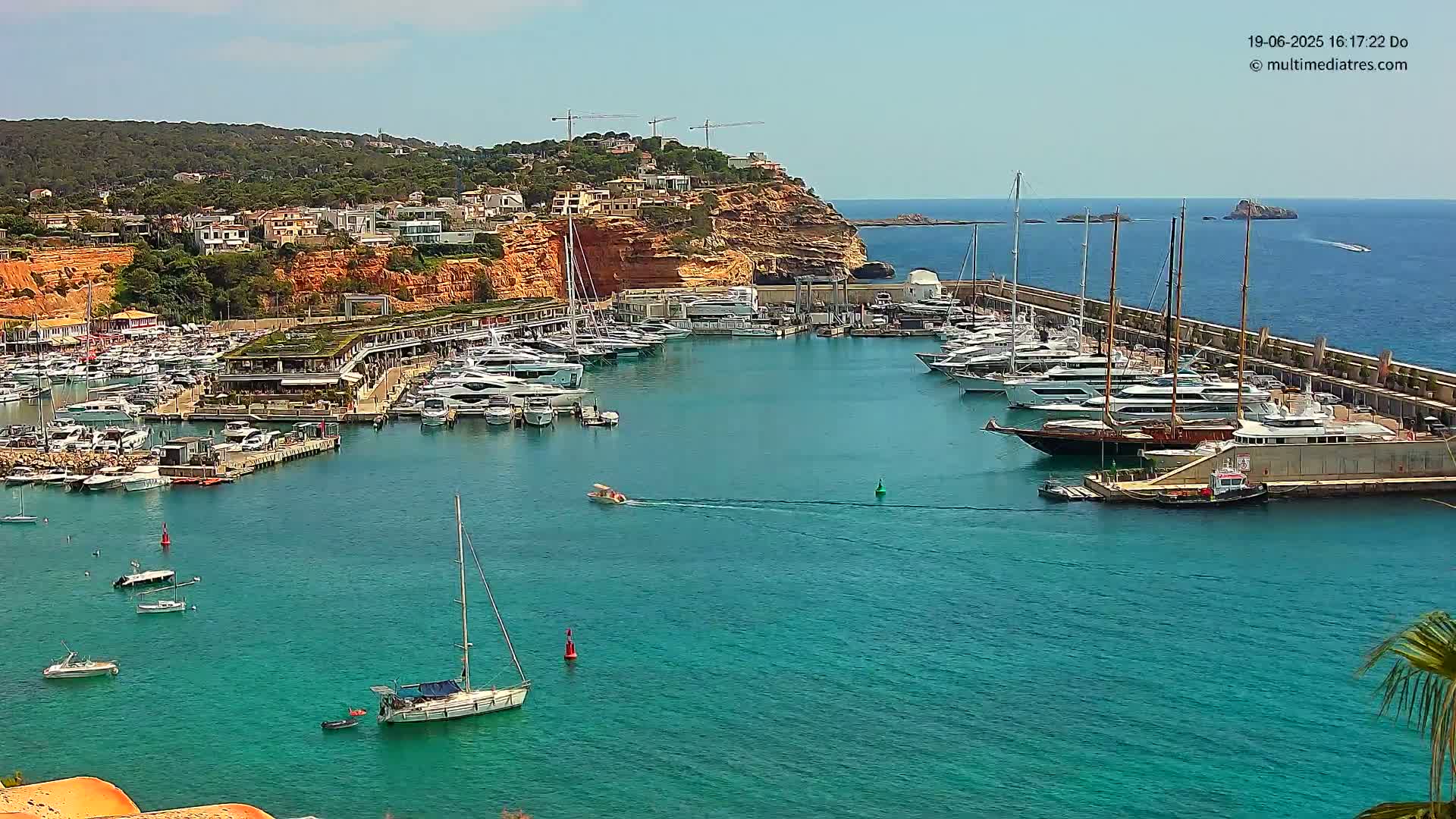 A harbor filled with numerous yachts and sailboats is nestled against a rocky, red-toned coastline under a sunny sky.