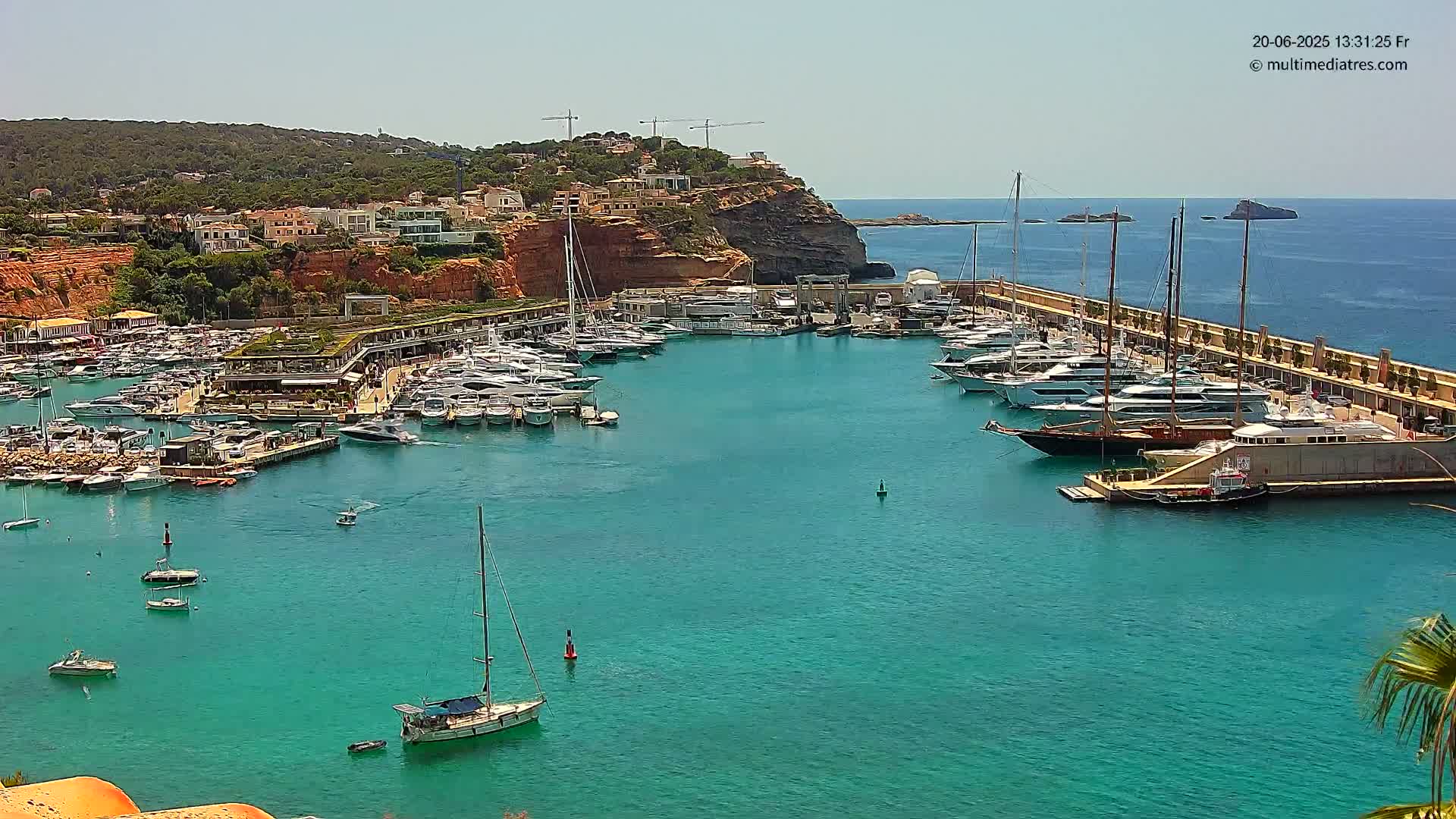 A marina filled with numerous boats and yachts is situated in a calm, turquoise bay, bordered by a red cliff topped with buildings and lush green vegetation under a clear, sunny sky.