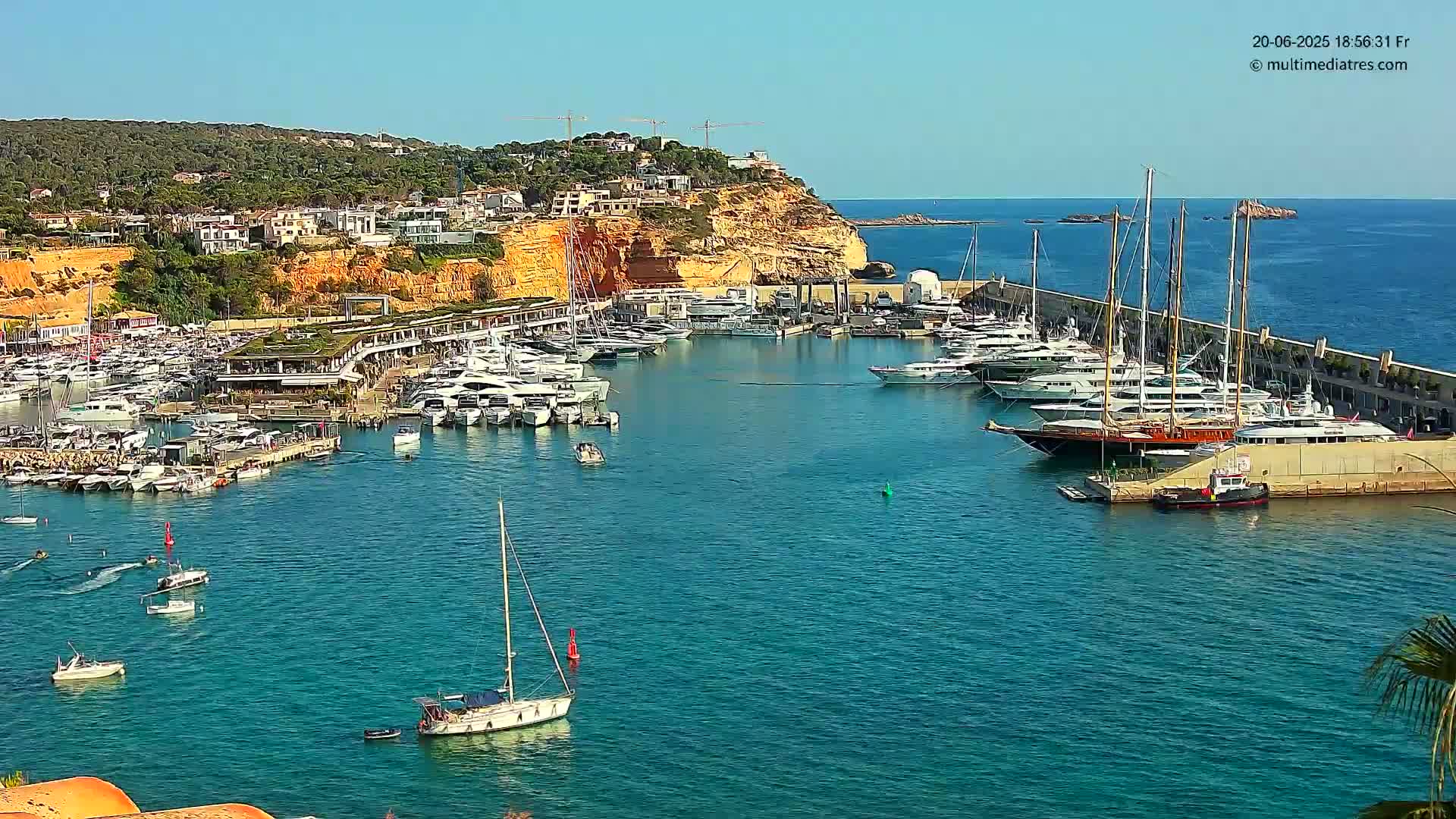 A marina filled with numerous yachts and boats is situated on calm, clear blue water under a sunny sky, nestled against a hillside town.