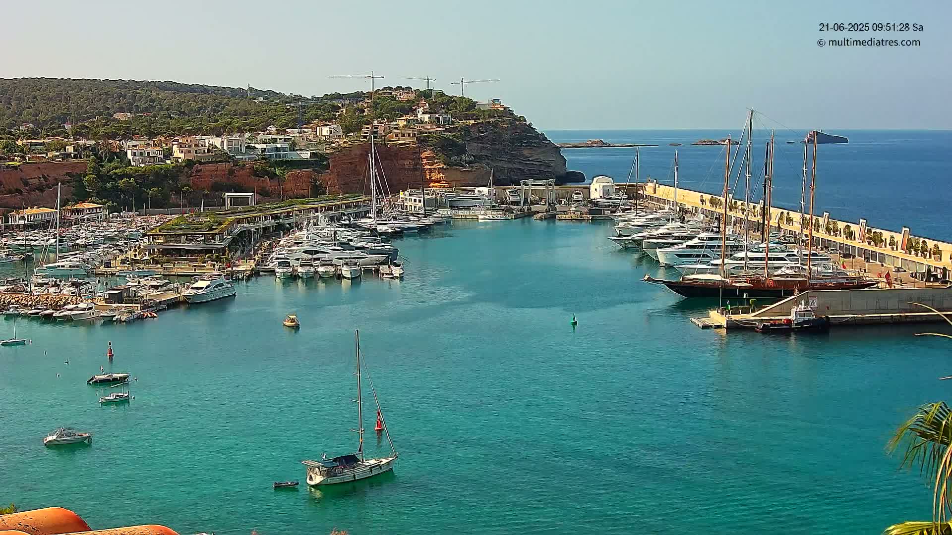 A marina filled with numerous yachts and sailboats is nestled against a hillside of buildings under sunny skies.