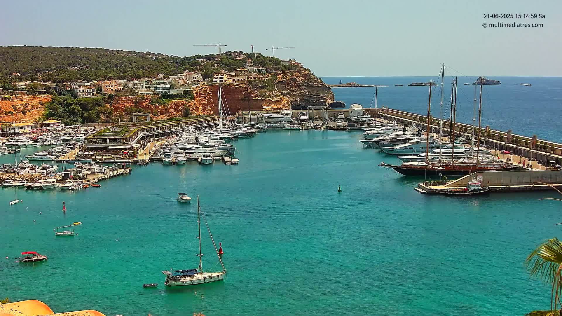 A marina filled with yachts and boats sits on a turquoise body of water next to a town built into a red cliff on a sunny day.