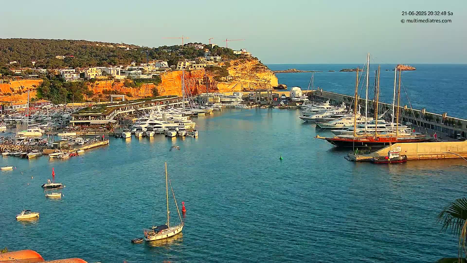 A calm, sunny day reveals a harbor filled with numerous yachts and sailboats docked along a cliffside, with additional boats in the water.