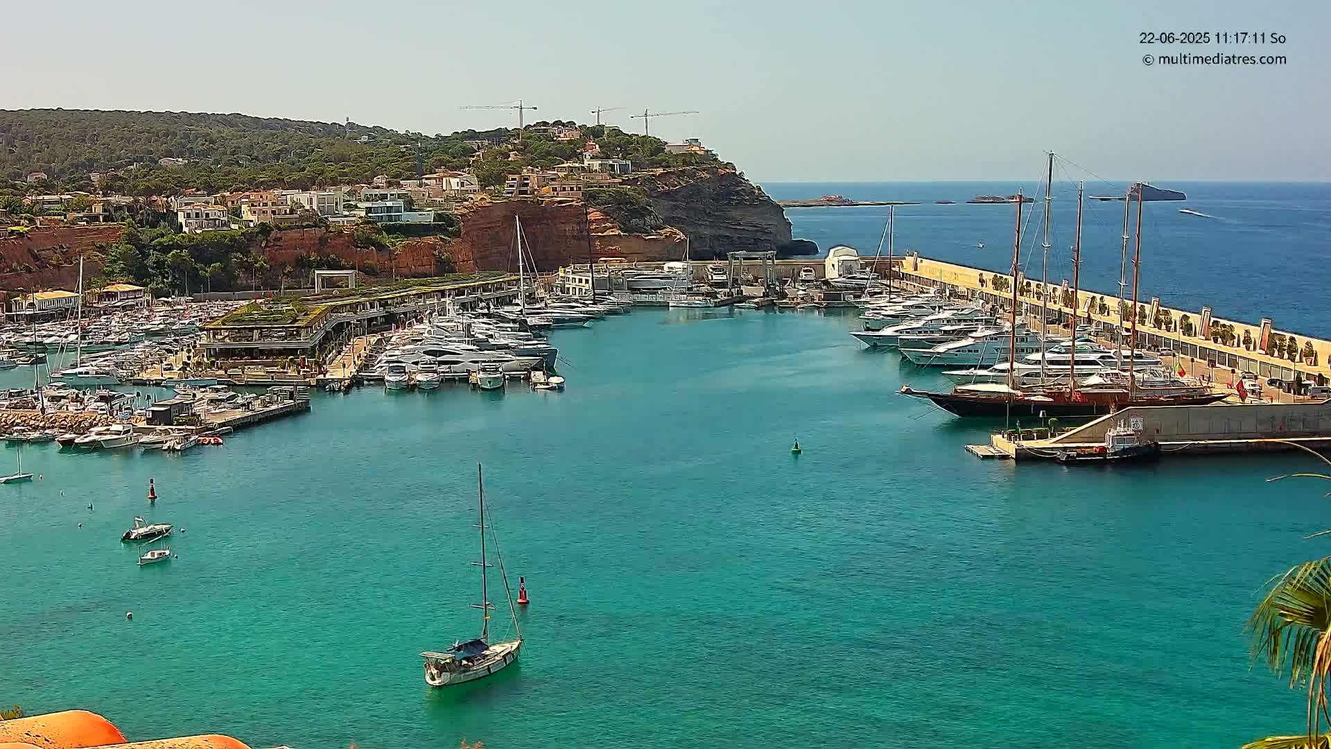 A sunny day reveals a harbor filled with numerous yachts and sailboats, nestled against a cliffside town.