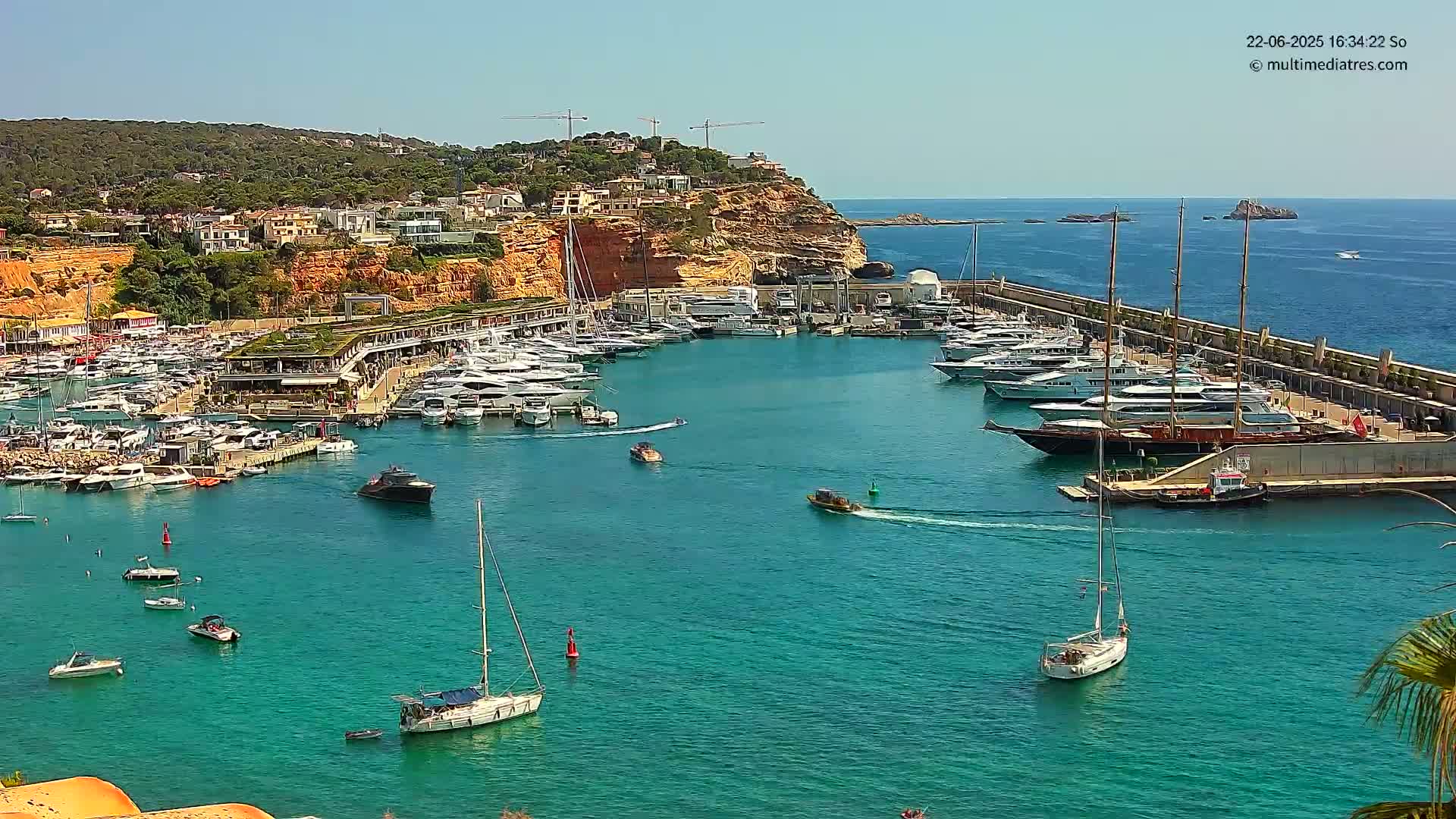 A marina filled with numerous yachts and sailboats is nestled against a rocky hillside under a clear, sunny sky.