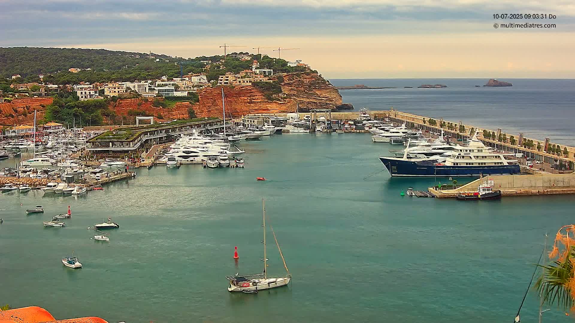 A harbor filled with numerous yachts and sailboats is nestled against a red cliffside, under a mostly cloudy sky.