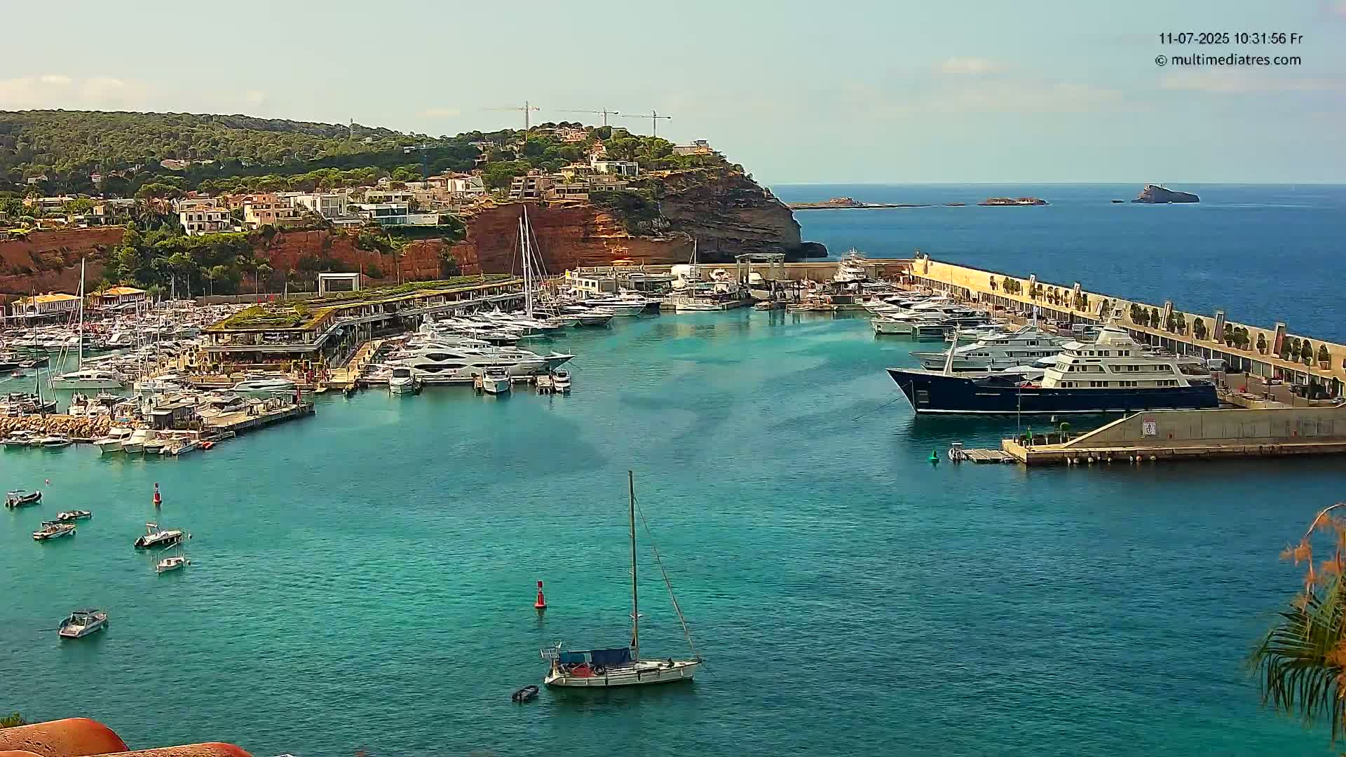 A marina filled with yachts and sailboats is nestled in a calm, turquoise bay, backed by a hillside town under a clear sky.