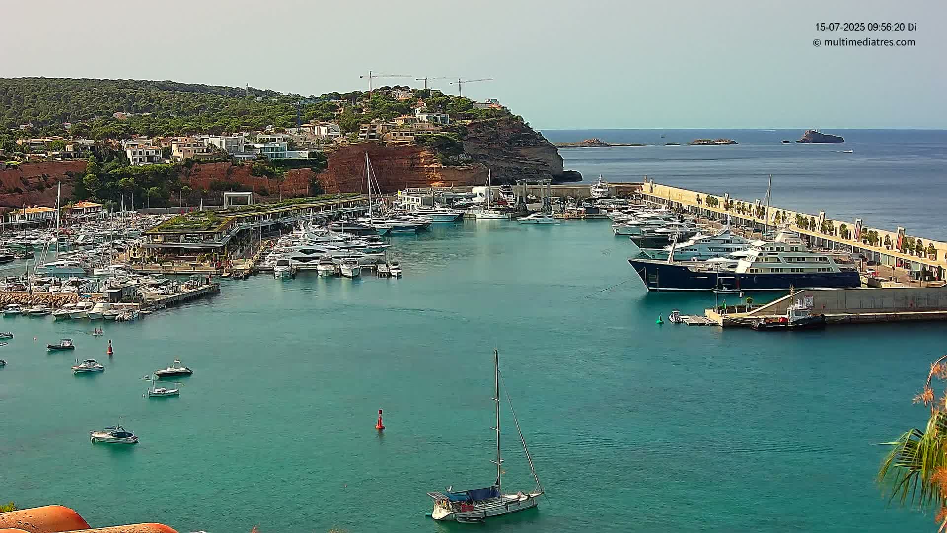 A marina filled with numerous yachts and boats is situated on a calm, teal-colored body of water, with a hillside of buildings visible in the background under a clear sky.