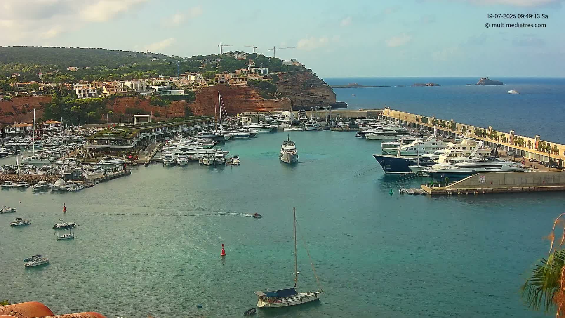 A marina filled with yachts and boats is nestled along a red cliffside overlooking a calm, blue sea under a mostly sunny sky.