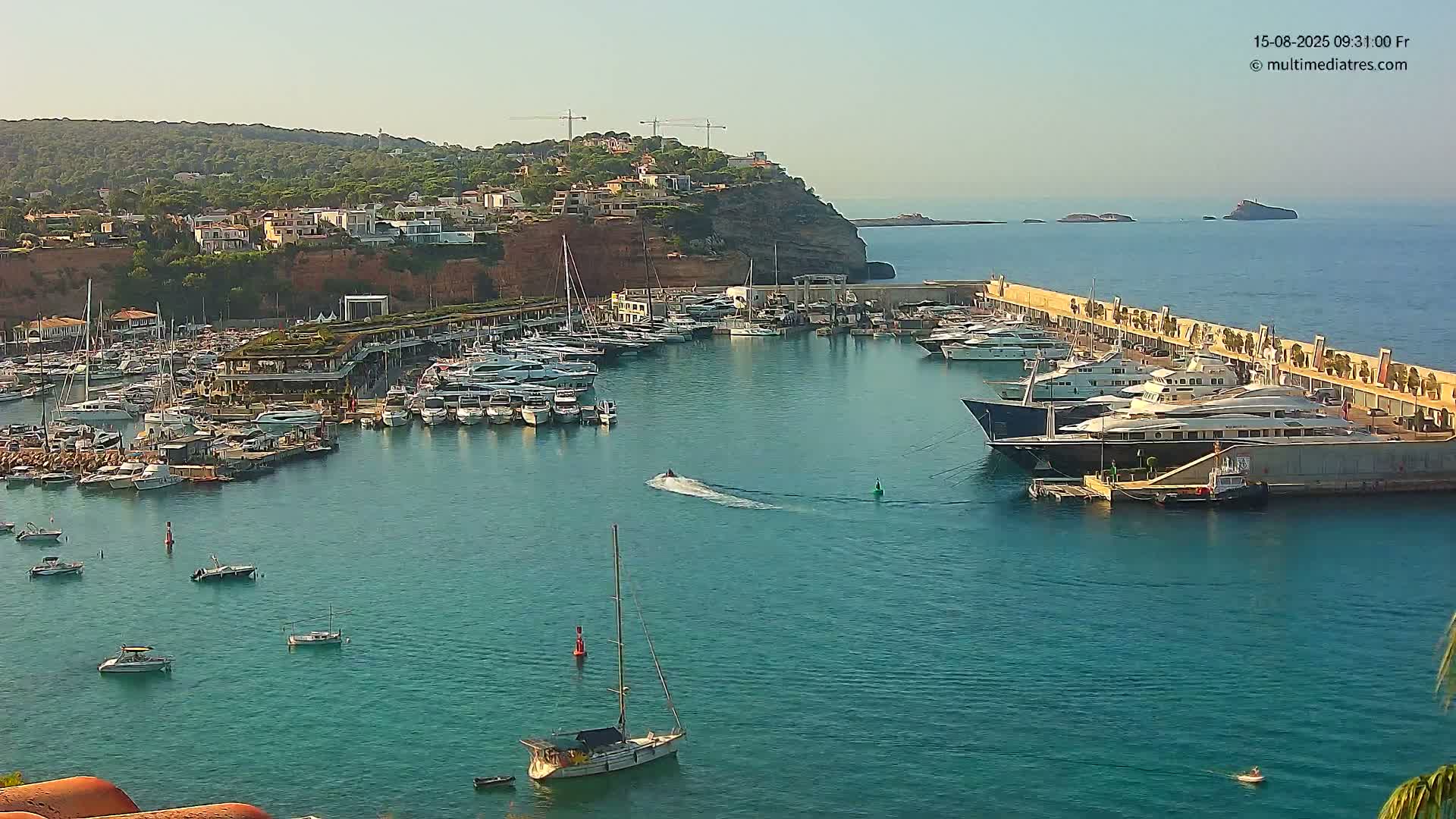 A marina filled with numerous yachts and boats is situated in a calm, clear body of water under a bright, sunny sky, with a hillside of buildings and green vegetation visible in the background.