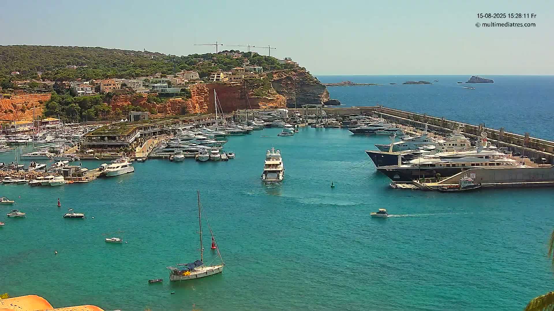 A marina filled with numerous yachts and boats is situated in a calm, clear body of water under a bright, sunny sky, with a hillside of buildings and green vegetation visible in the background.