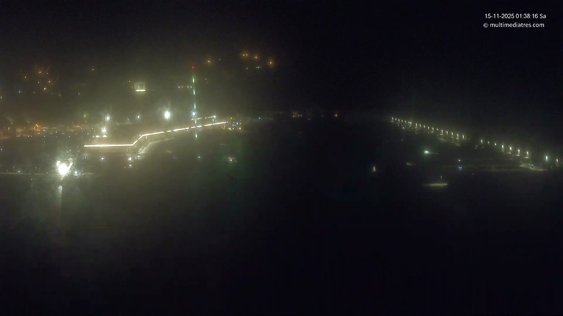 An expansive view of an urban or industrial area at night, heavily shrouded in fog or mist, with numerous bright lights reflecting on a damp surface and a long row of streetlights visible on the right.
