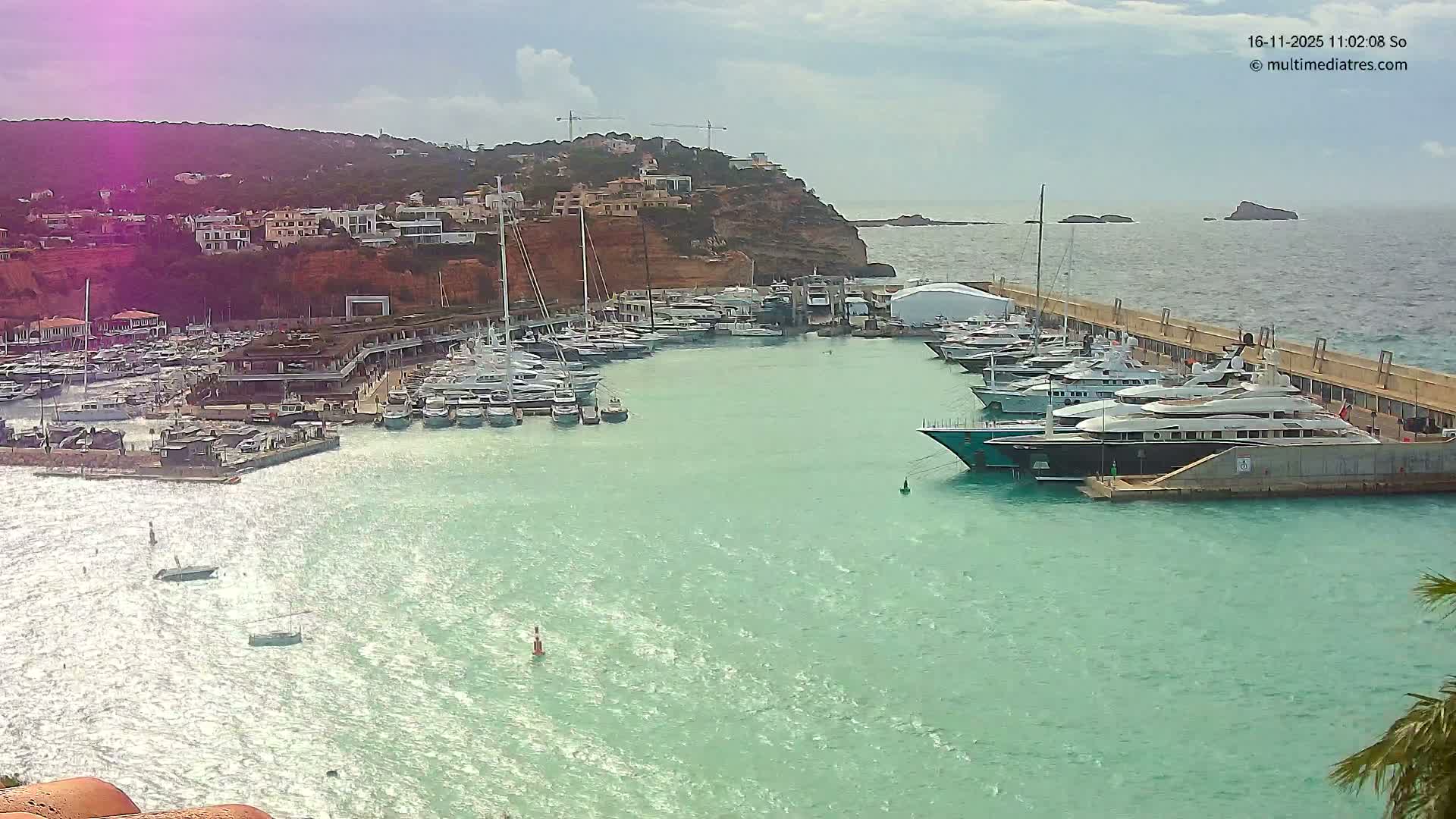 A bustling marina filled with numerous yachts and smaller boats floats on sparkling turquoise waters, set against a steep, building-covered hillside under a partly cloudy and breezy sky, with rocky islets visible in the distance.
