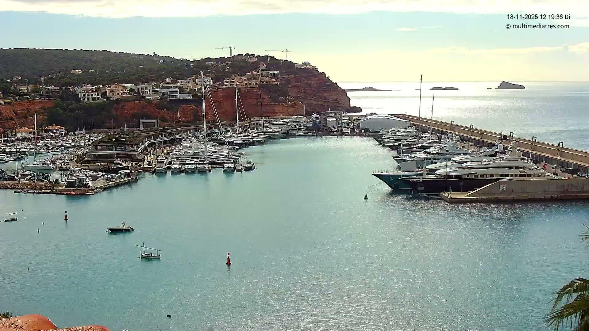 A bustling marina filled with numerous yachts and smaller boats floats on sparkling turquoise waters, set against a steep, building-covered hillside under a partly cloudy and breezy sky, with rocky islets visible in the distance.