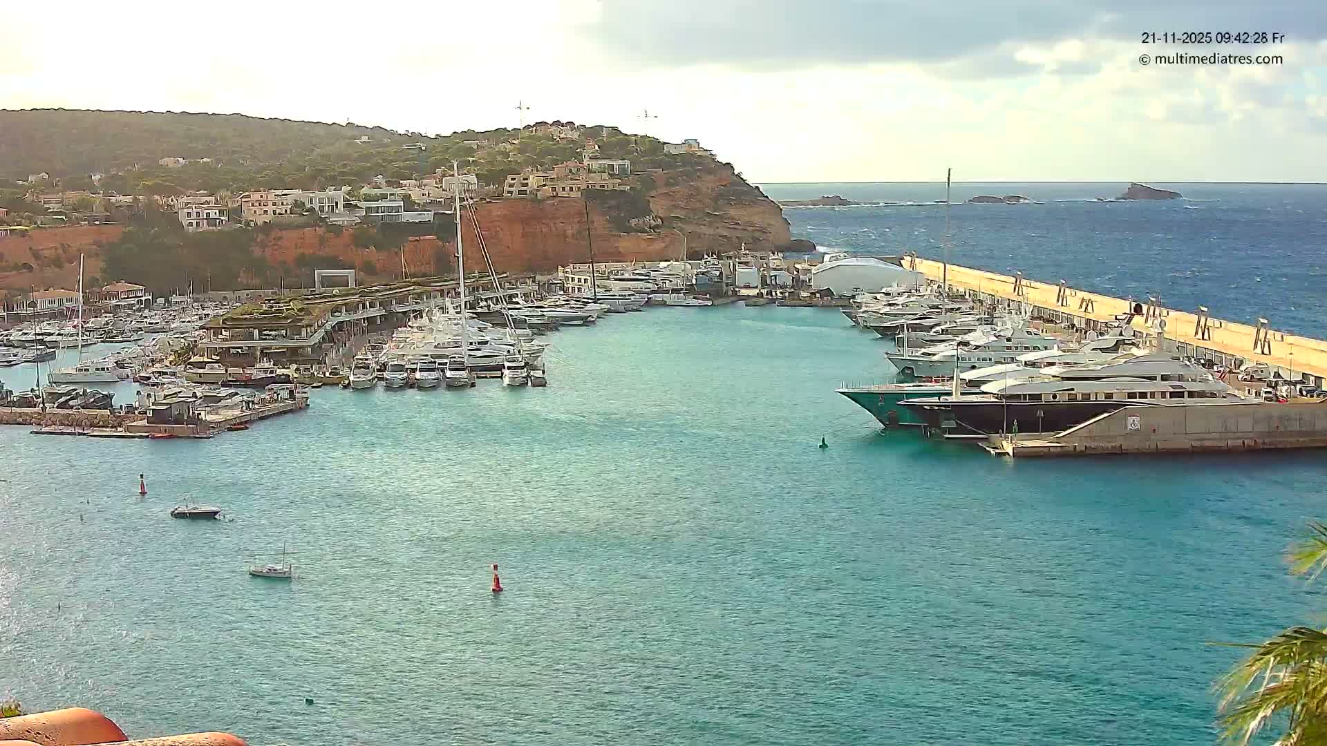 A large, busy marina filled with numerous yachts and boats is overlooked by a hillside covered in white buildings and greenery, all under a partly cloudy sky with noticeable waves in the open sea beyond the harbor.