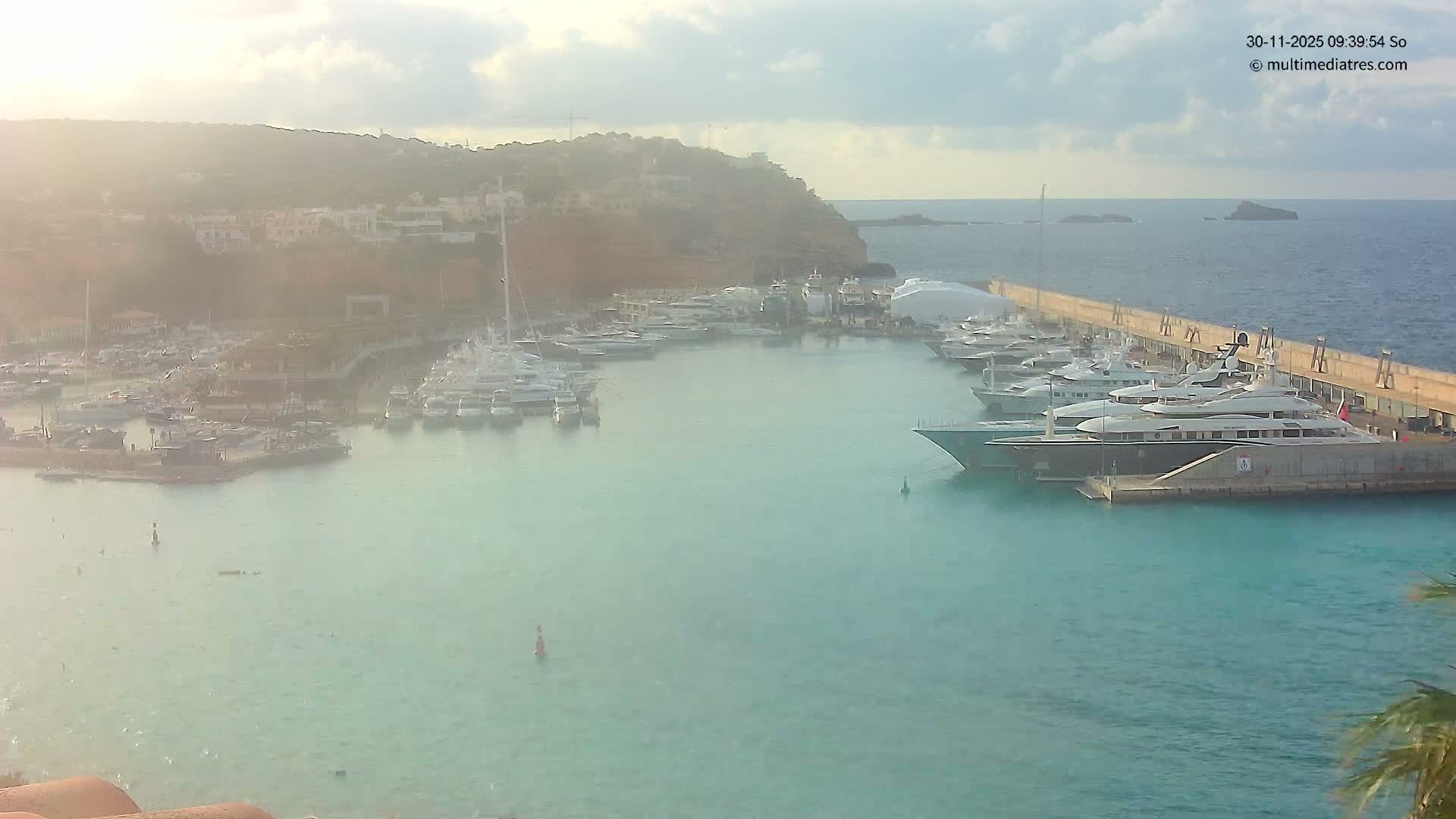 A bustling marina is seen filled with numerous yachts and boats docked along piers and in the clear blue water, all set against a hilly coastline dotted with buildings under a bright, hazy, and partly cloudy sky.
