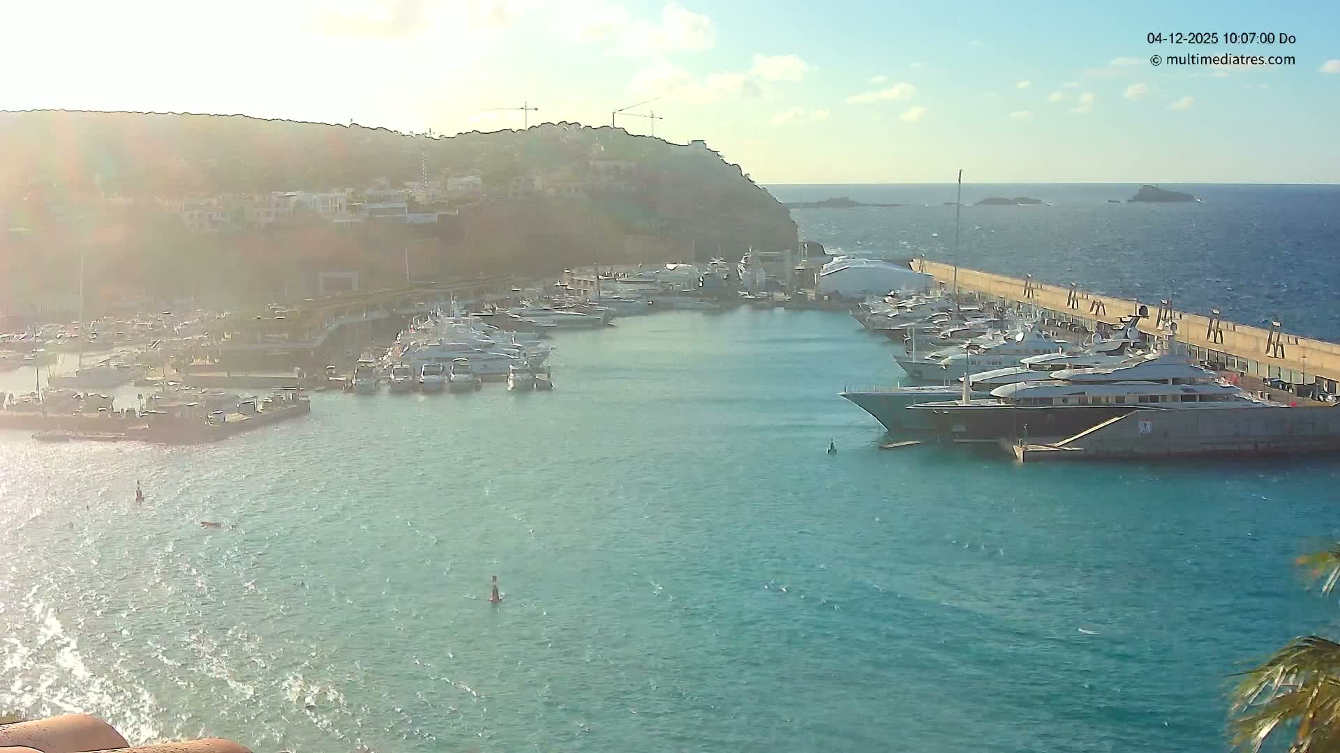 On a bright and sunny day, a bustling harbor with rippling turquoise waters is filled with numerous yachts and boats, bordered by a developed hillside with visible construction and a long pier extending to the clear open sea with distant islands.
