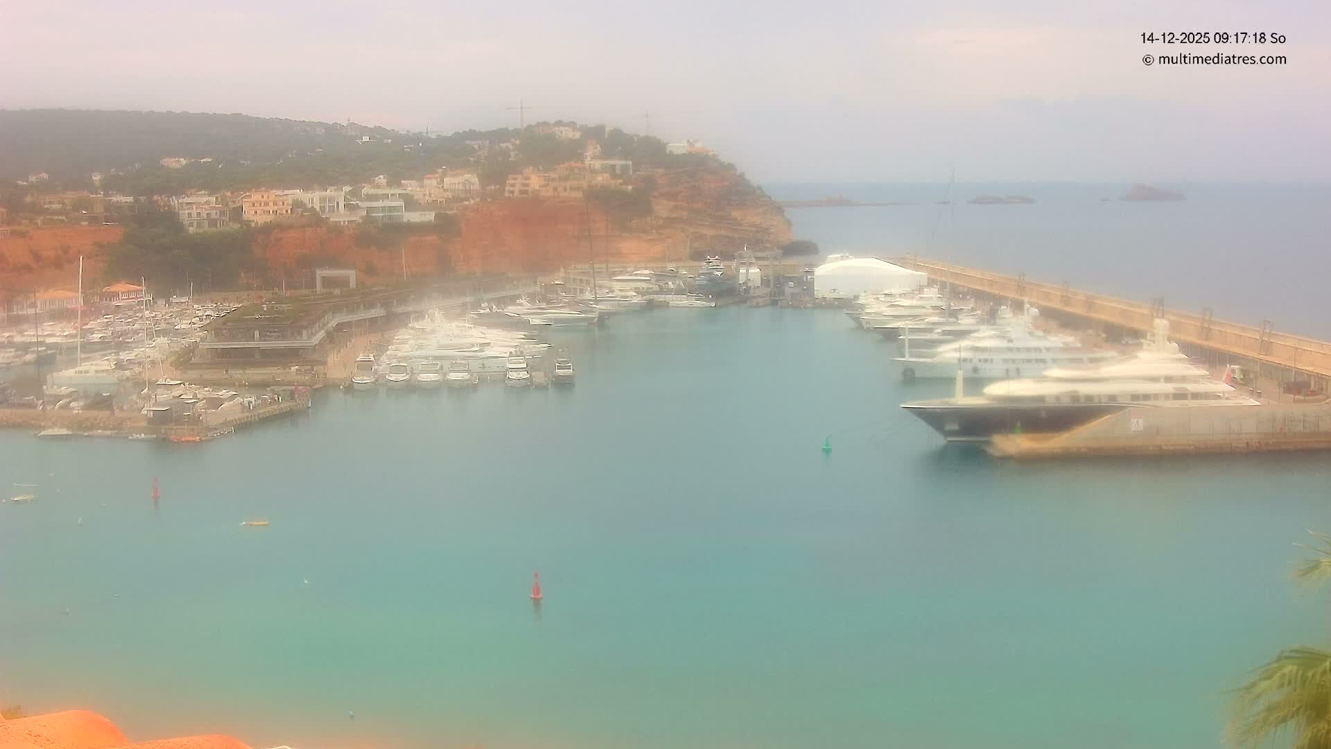 On a bright and sunny day, a bustling harbor with rippling turquoise waters is filled with numerous yachts and boats, bordered by a developed hillside with visible construction and a long pier extending to the clear open sea with distant islands.