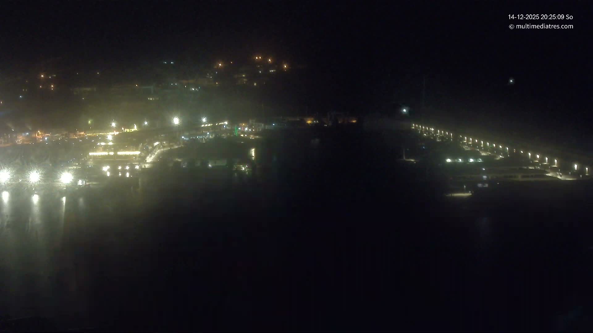 On a bright and sunny day, a bustling harbor with rippling turquoise waters is filled with numerous yachts and boats, bordered by a developed hillside with visible construction and a long pier extending to the clear open sea with distant islands.