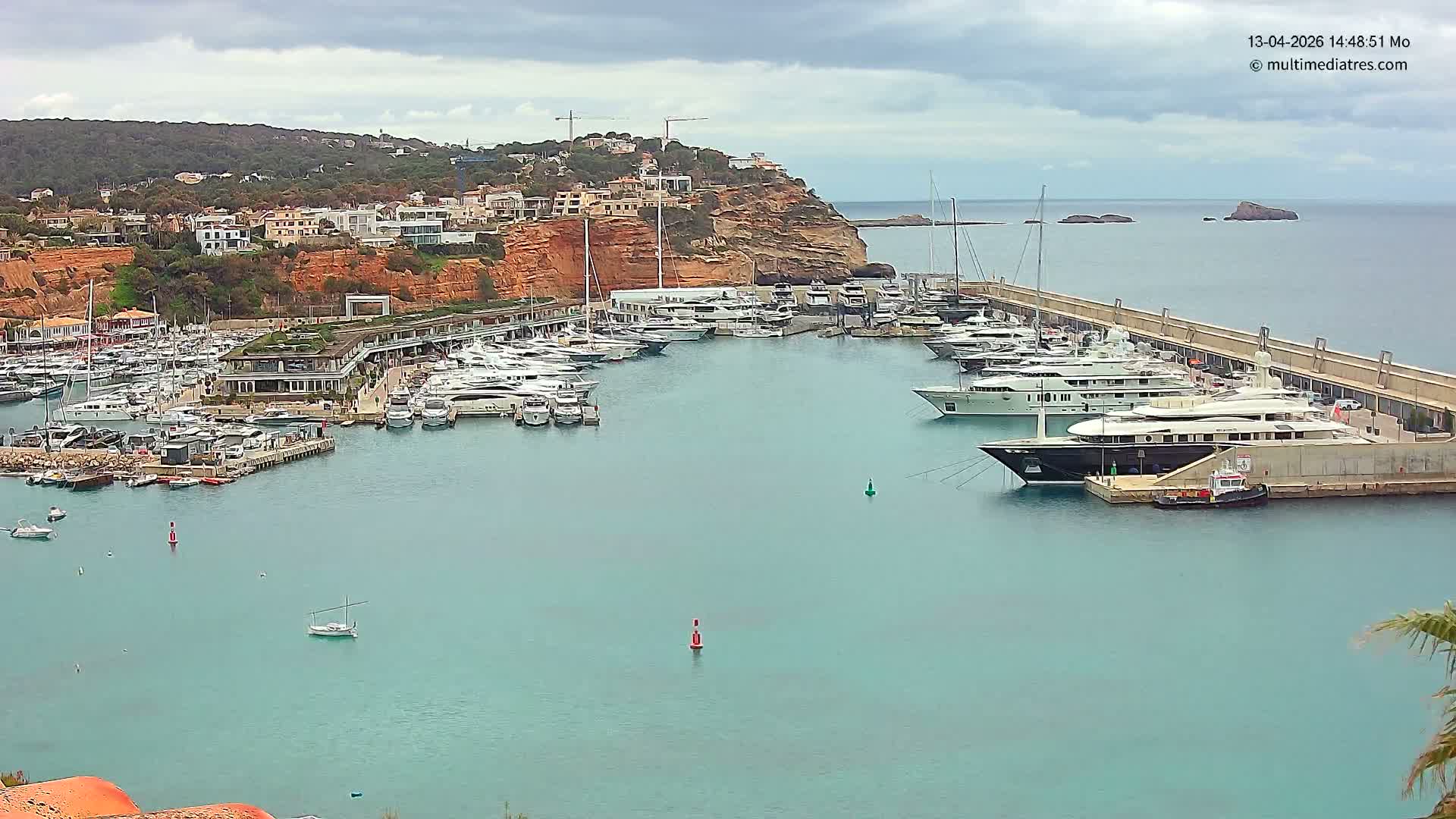 On a bright and sunny day, a bustling harbor with rippling turquoise waters is filled with numerous yachts and boats, bordered by a developed hillside with visible construction and a long pier extending to the clear open sea with distant islands.