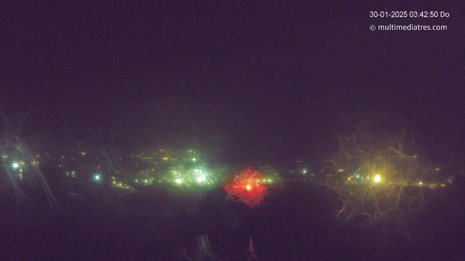 A nighttime long shot shows a distant town illuminated by scattered lights, with fireworks exploding in the center and to the right.