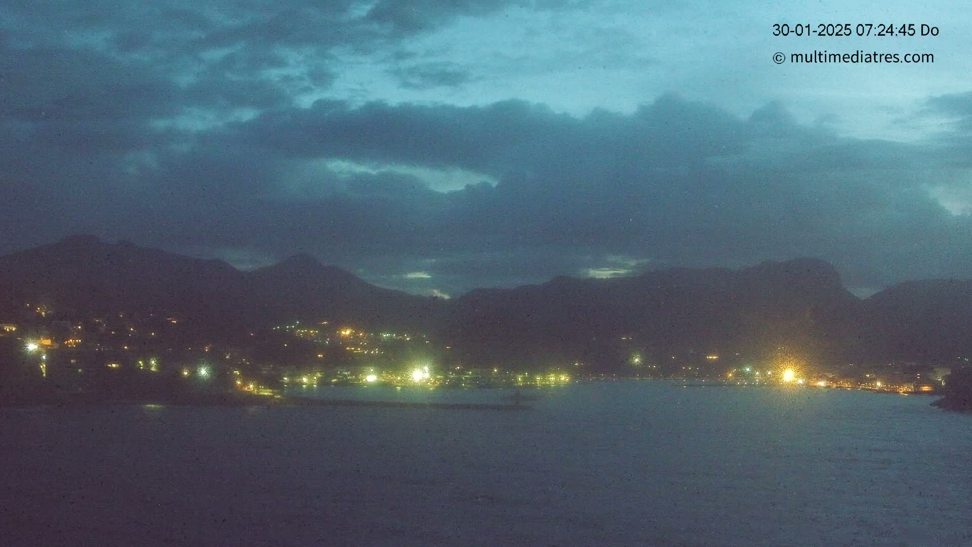 A nighttime view of a coastal town nestled between dark mountains under a cloudy, dusky sky.