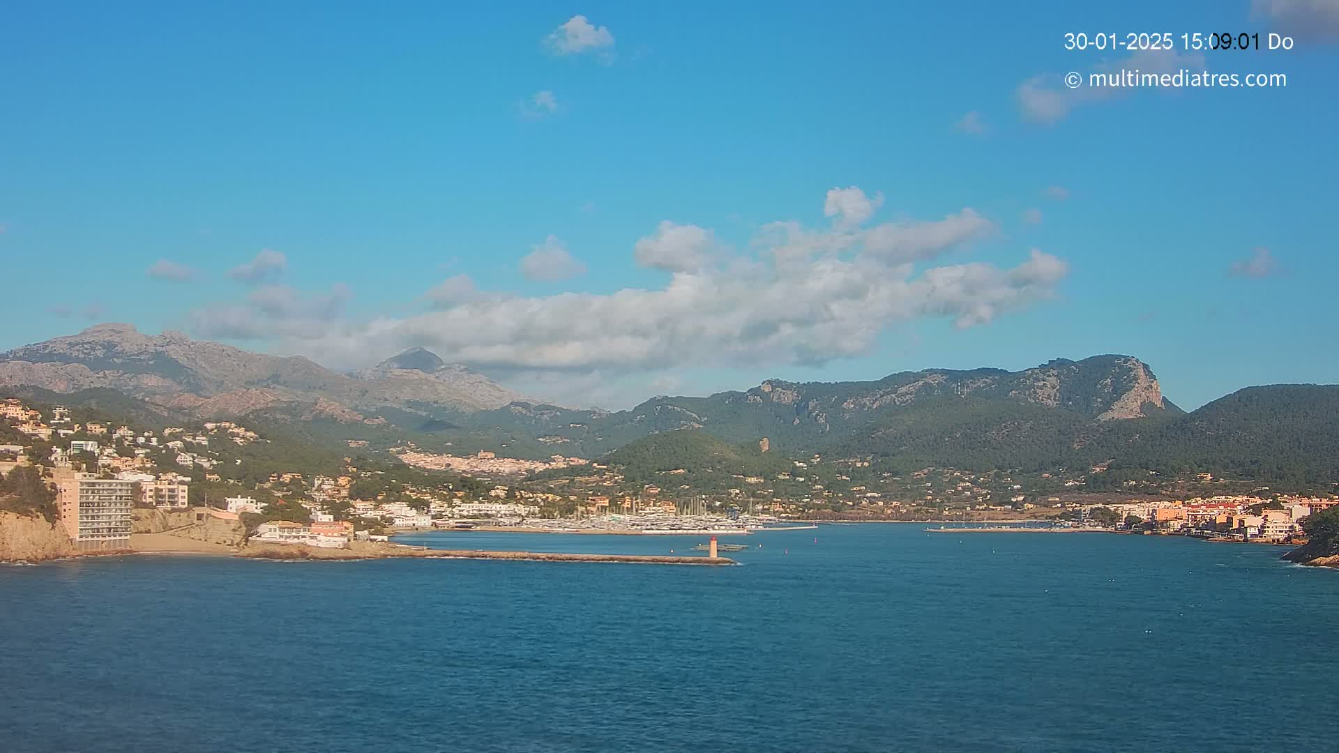 A coastal town nestled against a mountain range under a mostly clear blue sky with some scattered clouds, features a harbor with numerous boats.
