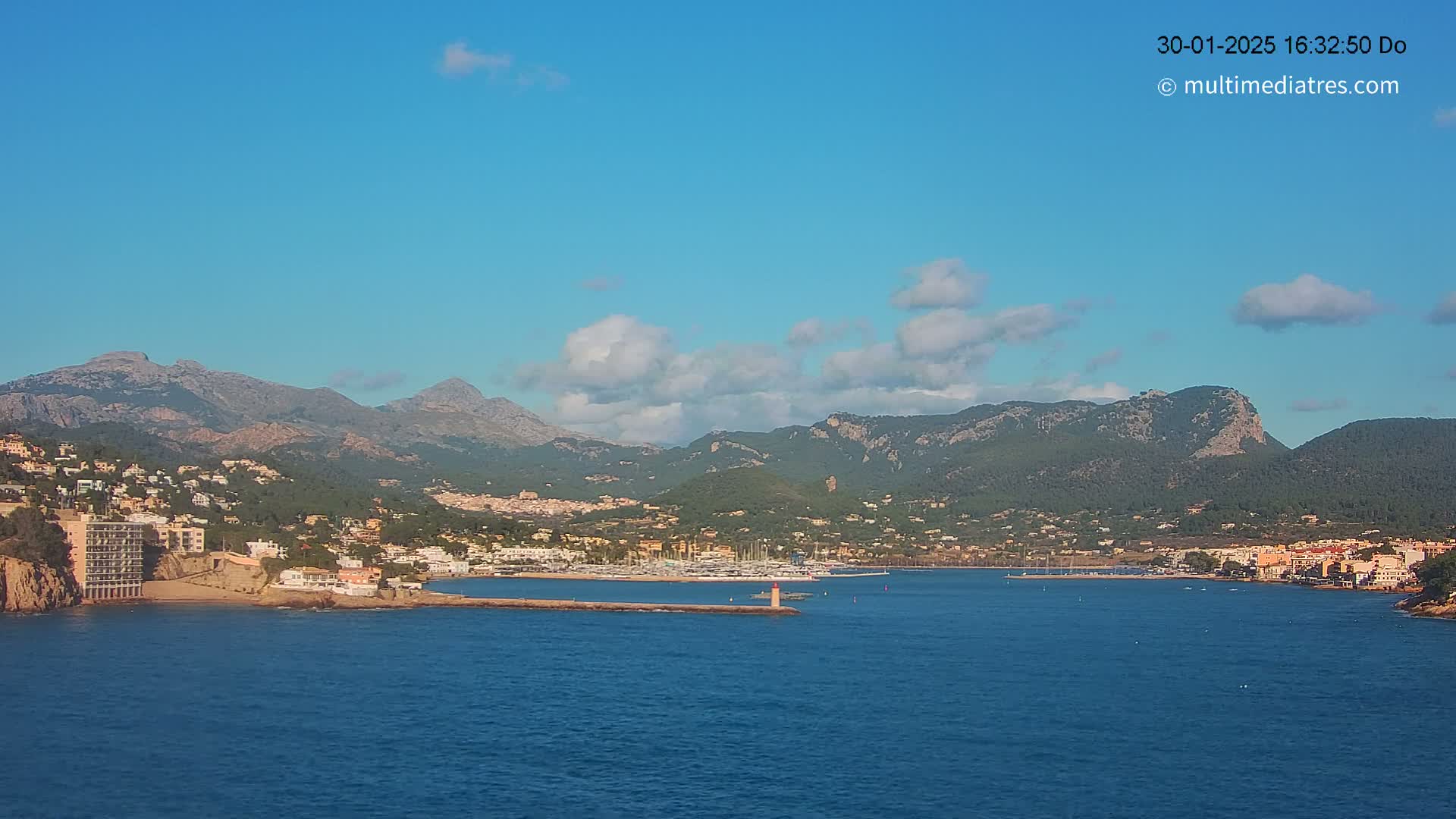 A coastal town nestled between mountains under a mostly clear blue sky with a few scattered clouds, features a harbor with boats and a breakwater.