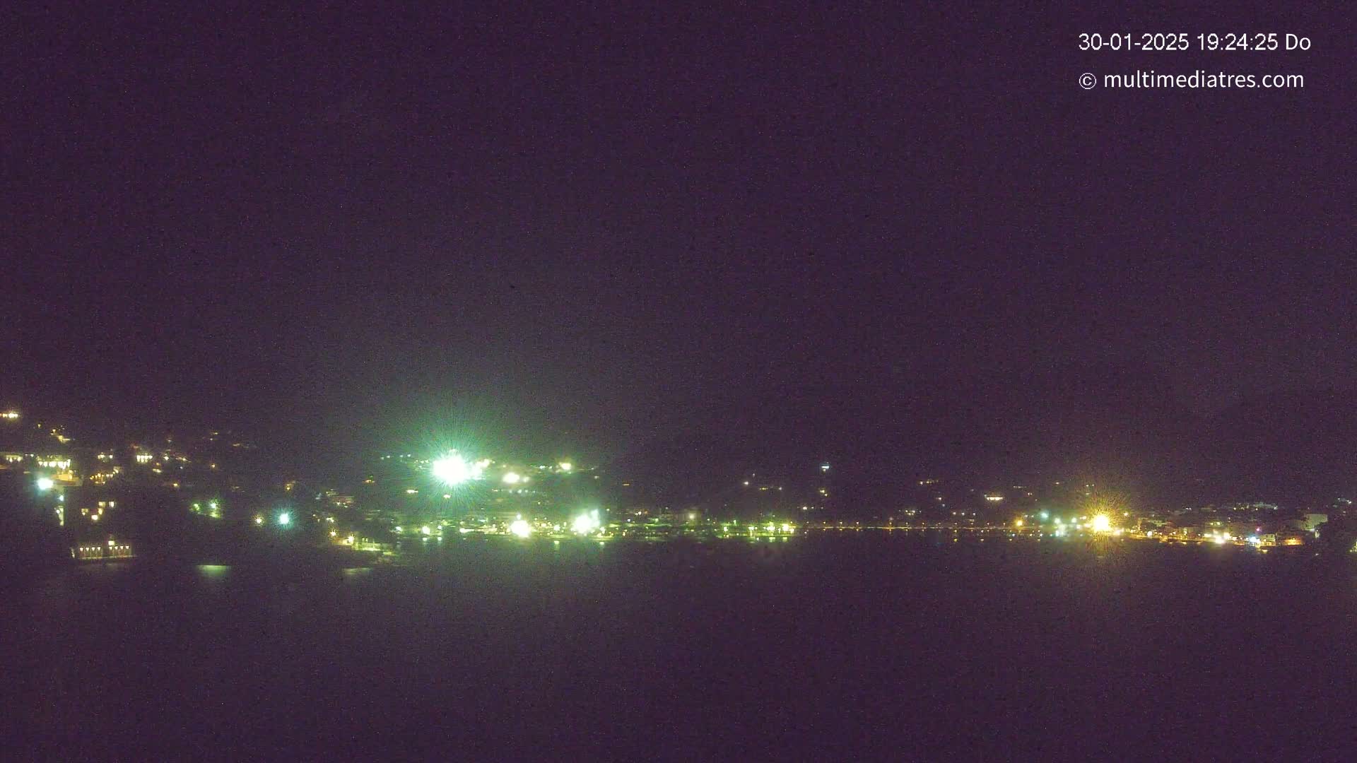 A nighttime long shot shows a coastal town with scattered lights along a dark body of water under a clear, dark sky.