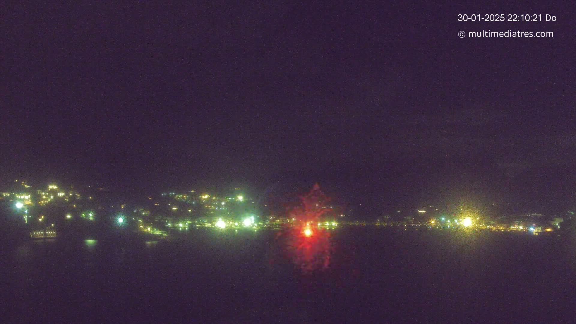 A nighttime, coastal scene shows a red firework exploding over a dark body of water in front of a city with illuminated buildings on a hill.