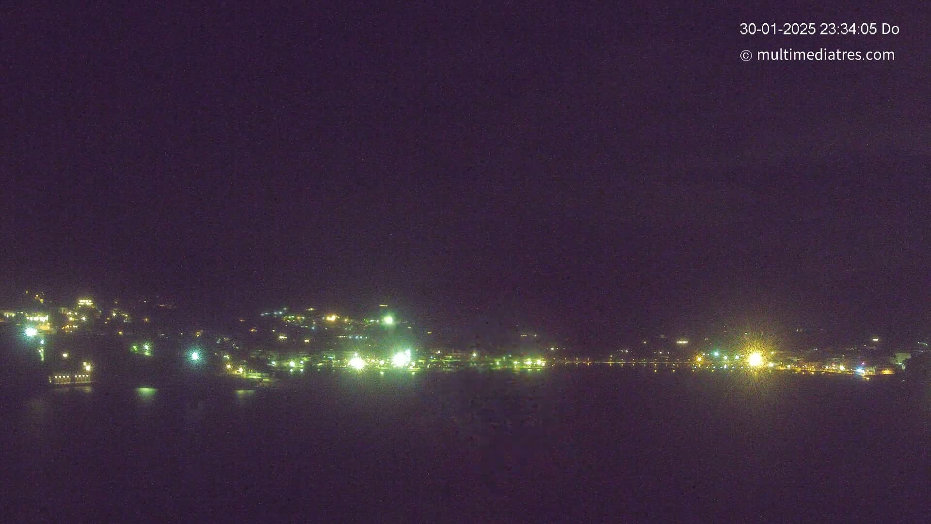 A nighttime long shot shows a dark body of water reflecting the lights of a town situated on a hillside under a clear, dark sky.