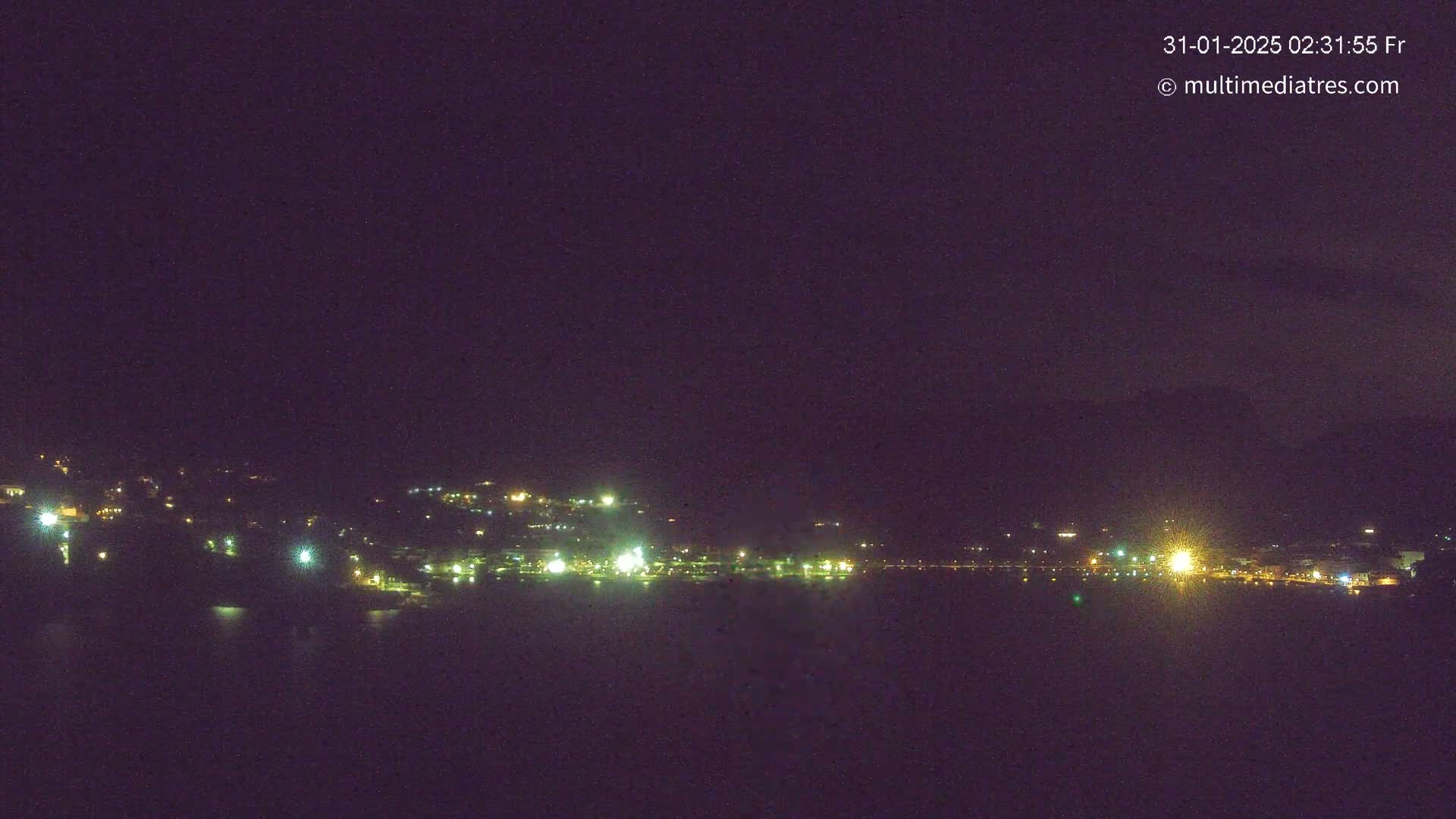 A nighttime long shot shows a dark body of water reflecting the faint lights of a town nestled on a hillside under a clear, dark sky.