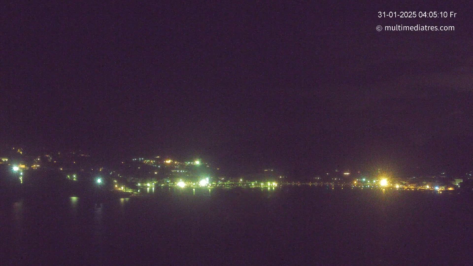 A nighttime long shot of a dark, calm body of water reflecting the lights of a town nestled on a hillside under a clear, dark sky.