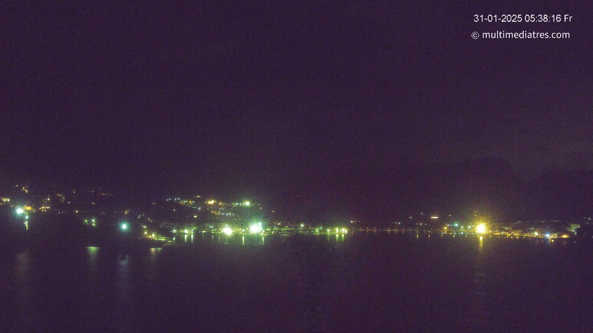 A nighttime view shows a town's lights across a dark body of water under a clear, dark sky.