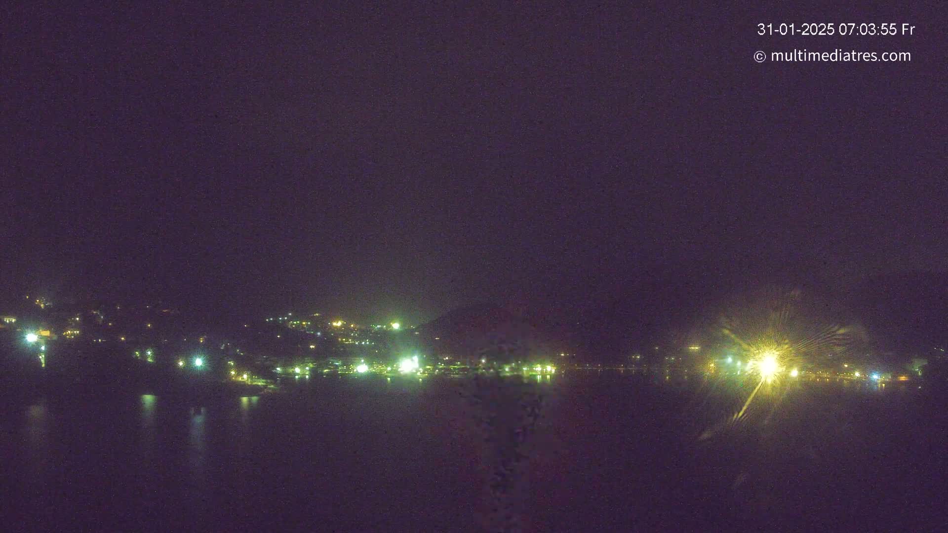 A nighttime view shows a dark body of water reflecting the lights of a town nestled against a hillside under a clear, dark sky.