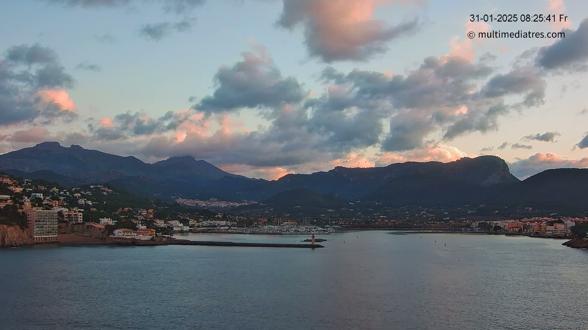A coastal town nestled between mountains under a partly cloudy sunset sky.