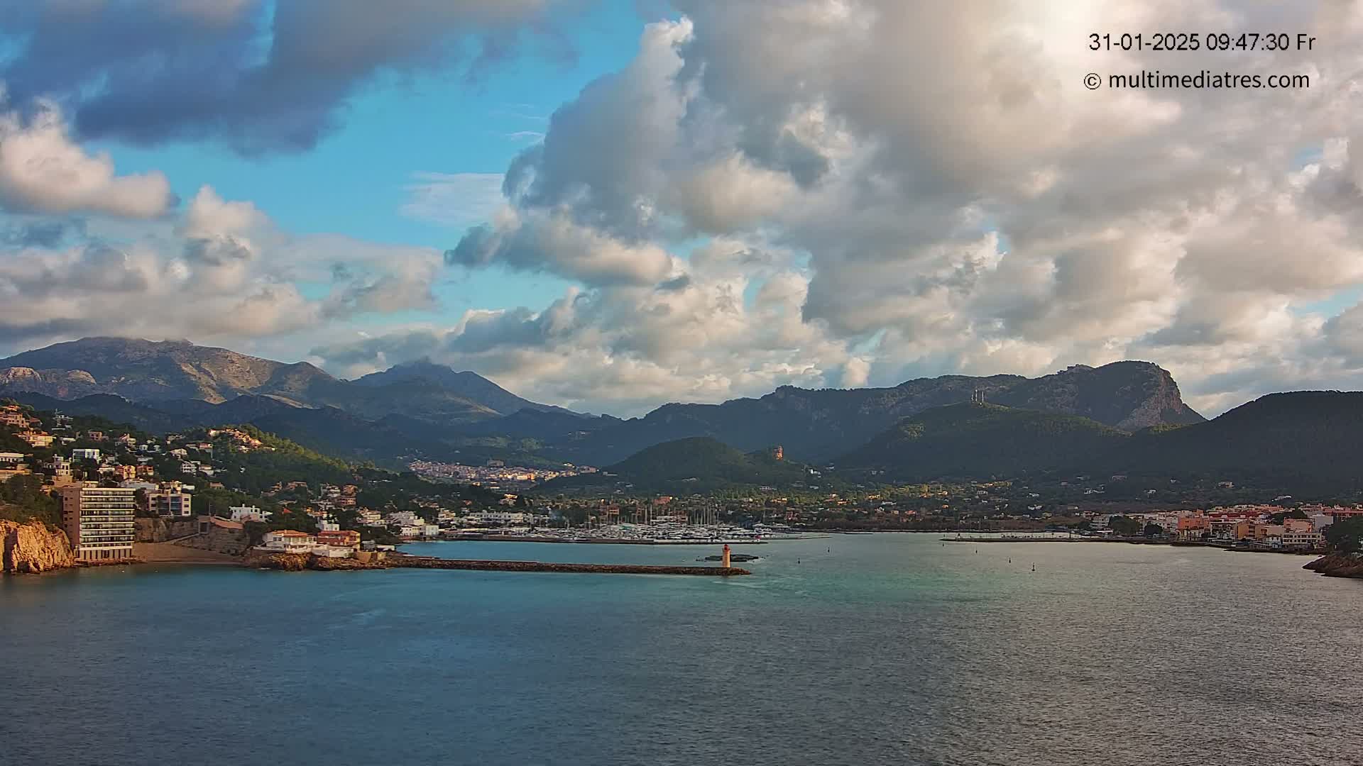 A coastal town nestled at the base of mountains under a partly cloudy sky sits on a calm body of water with a harbor filled with boats.